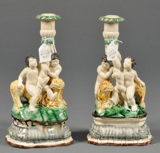 Pair of Creamware Figural Candlesticks, Continenta: Pair of Creamware Figural Candlesticks, Continental, 19th century, each modeled with putti seated atop a lion and supporting a cornucopia-form sconce, all atop a raised base, translucent glazes, ht. 1