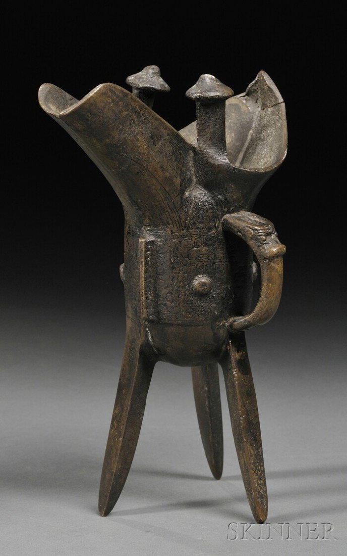 Bronze Jue, China, 19th century, raised on three s: Bronze Jue, China, 19th century, raised on three slender blade-shaped feet, the body cast with a taotie mask to each side, upright posts to each side of the mouth rim, ht. 7 1/4 in.
