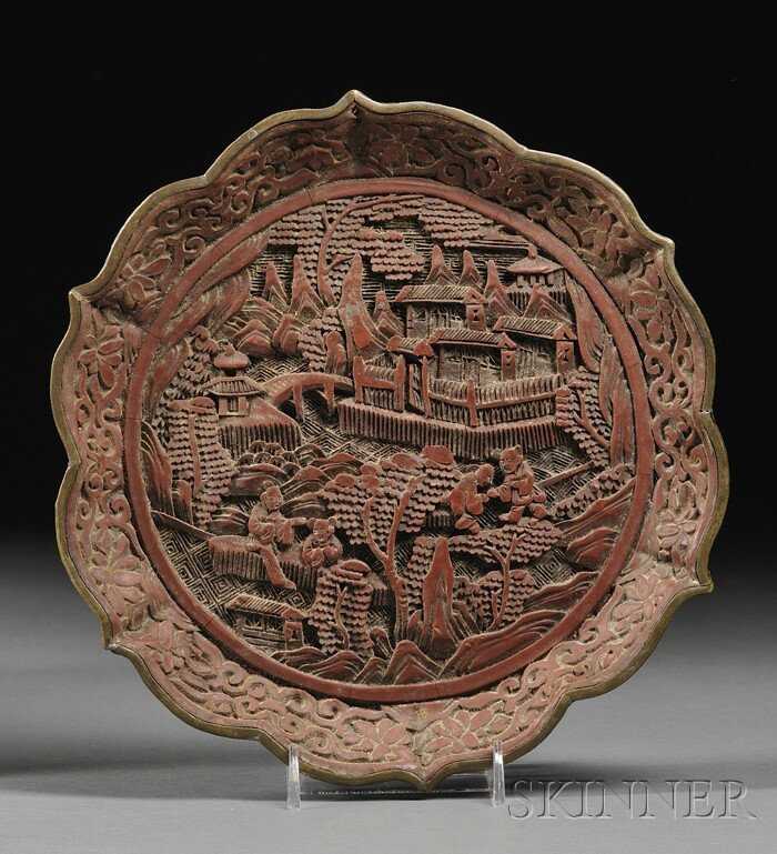 Cinnabar Plate, China, late 19th century, the inte: Cinnabar Plate, China, late 19th century, the interior of the petal-rimmed plate carved in relief with figures in a rustic landscape, encircled by flower borders, brocade design to the base and with a