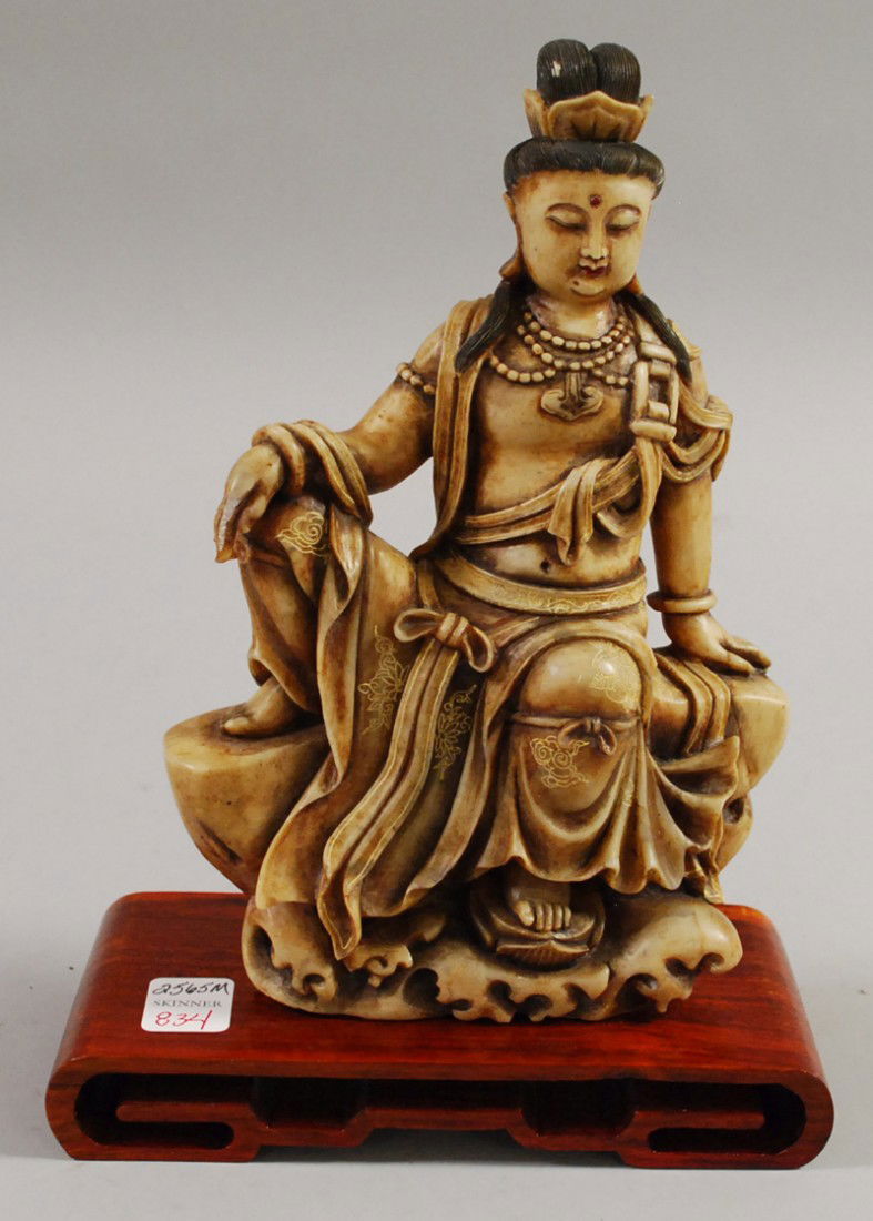 Soapstone Carving, China, carved as the seated fig: Soapstone Carving, China, carved as the seated figure of Guanyin in the royal ease pose atop a rockery, her left foot stepping on a lotus above crashing waves, wood stand, ht. 9 1/2 in.
