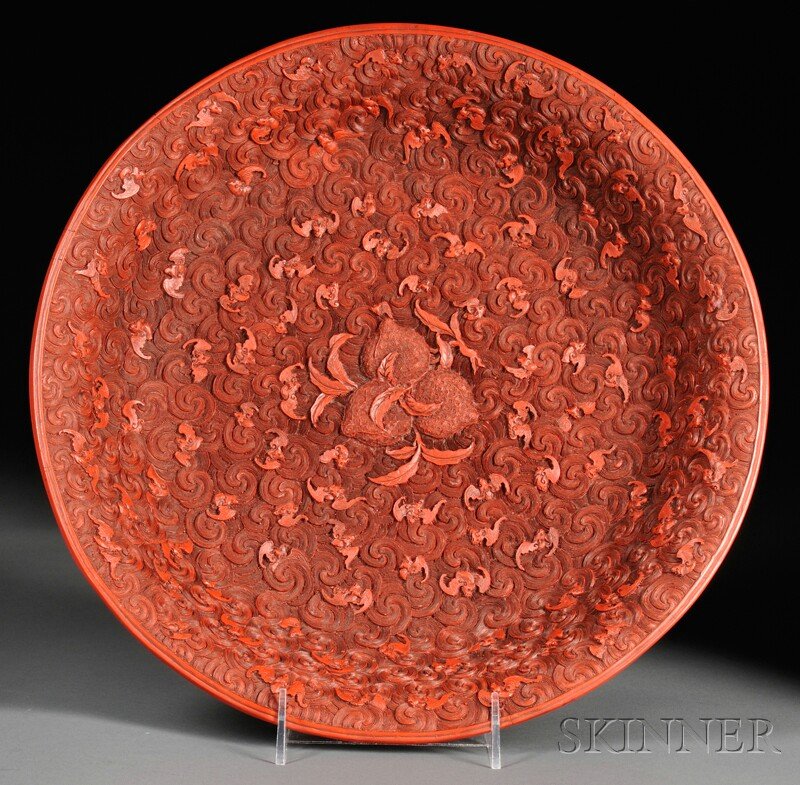 Faux Cinnabar Lacquer Plate, China, the interior i: Faux Cinnabar Lacquer Plate, China, the interior intricately carved with three peaches at the center and bats flying on a ground of rolling clouds, the exterior with peonies and blossoms on a geometri