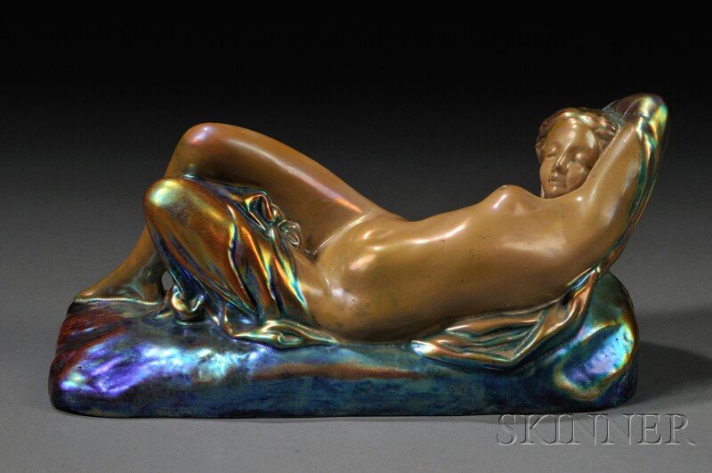 , Zsolnay Reclining Female Nude Figure, Art potter: Zsolnay Reclining Female Nude Figure Art pottery Hungary, c. 1912 Reclining on a rectangular plinth in iridescent glaze, round raised maker's mark, impressed 8577, some glaze imperfections, lg. 8 1/4 