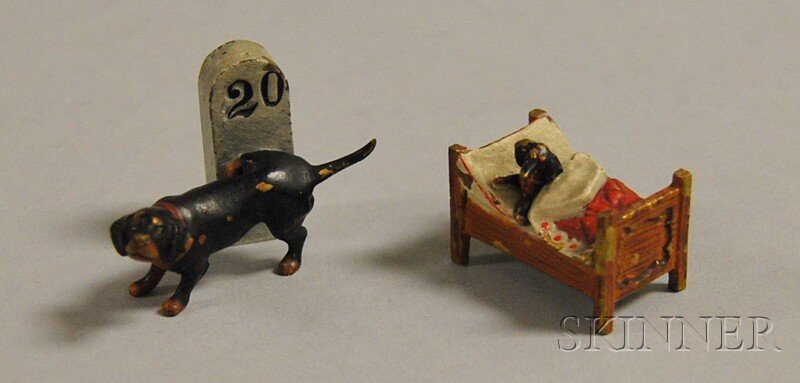 Two Austrian Miniature Cold-painted Bronze Dachsh: Two Austrian Miniature Cold-painted Bronze Dachshund Figurals, early 20th century, one urinating on a 20km road marker, and one in bed, original paint, ht. 1 1/8, lg. 1 1/8 in., respectively.