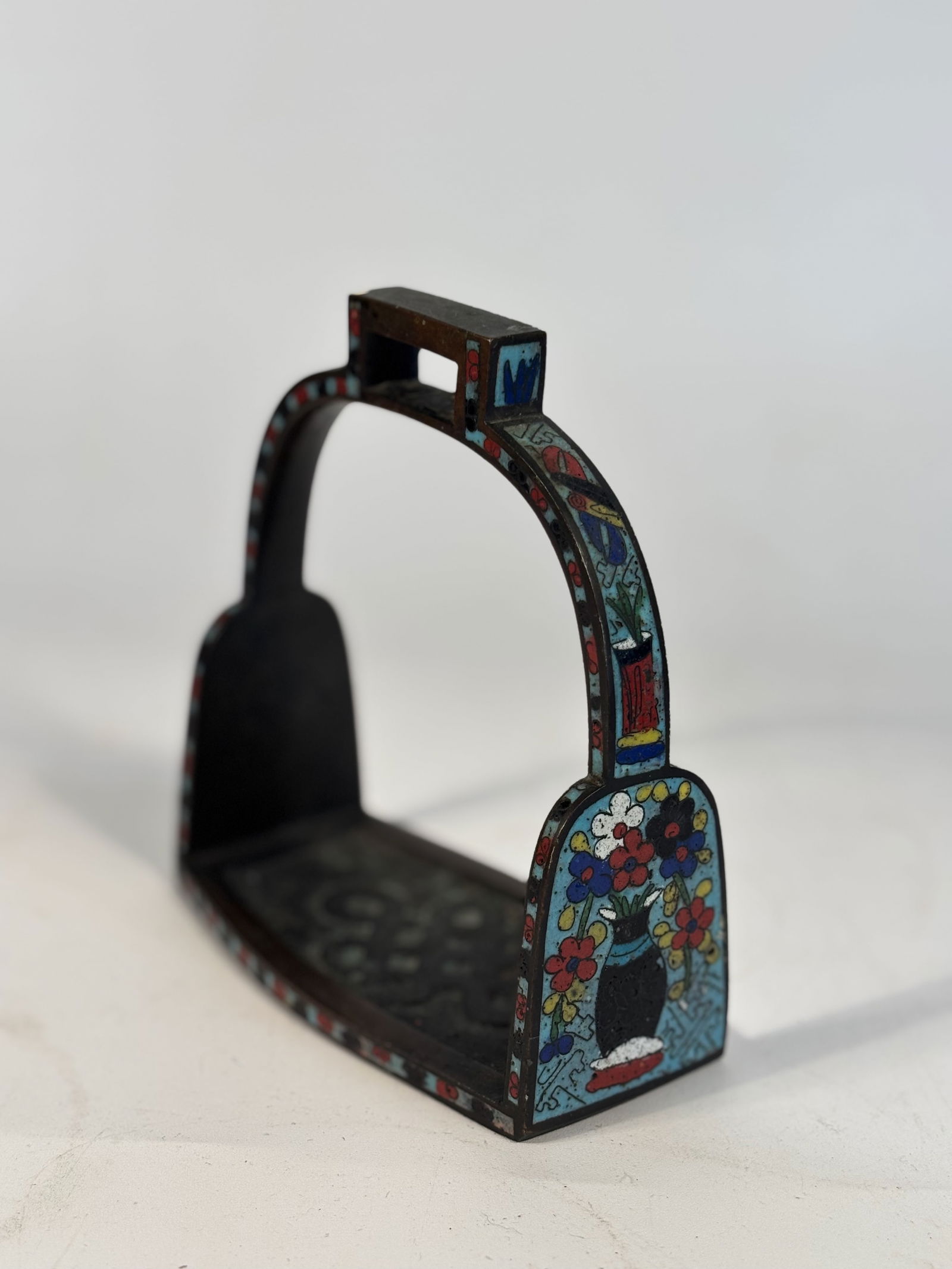 A Chinese Cloisonné Enamel Stirrup with Floral and Vase Motifs: Qing Dynasty (1644-1912); A finely enameled Chinese stirrup of classic arched form with cloisonné enamel decoration on both sides and the upper arch. Each side is decorated with a central black vase