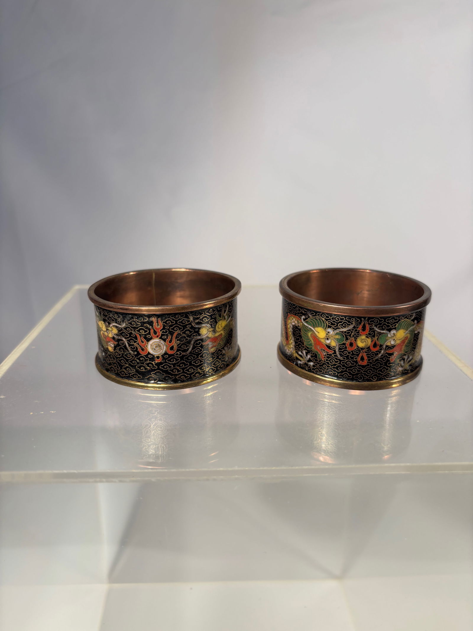 Pair Chinese Cloisonné Napkin Rings with Dragon Motifs: pair of Chinese cloisonné napkin rings, each decorated with intricate multicolored enamel designs on a black ground. The rings feature auspicious motifs, including imperial five-clawed dragons, flami