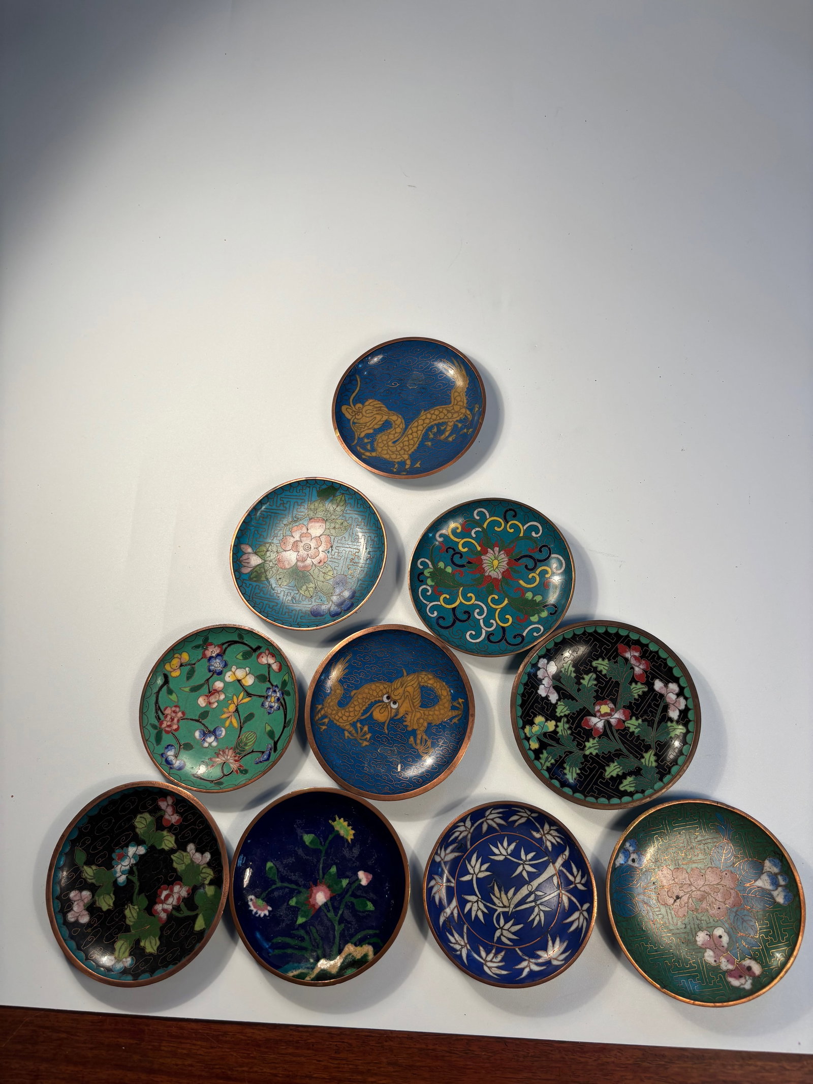 Set of Ten cloisonné plates: 19th century;A collection of small enamel dishes featuring intricate designs, including dragons, floral patterns, and geometric motifs. Diameter: