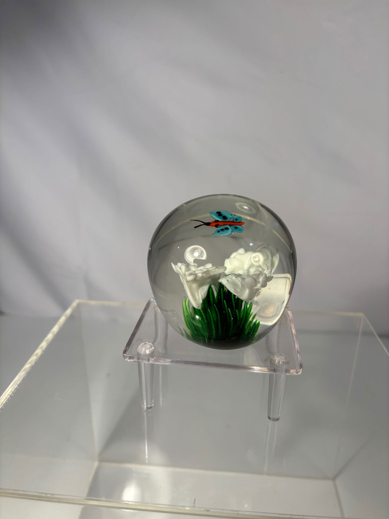 Art Glass Paperweight with Butterfly and White Flower: Charming Dynasty Gallery collectible glass paperweight, featuring a delicately crafted turquoise and red butterfly hovering above a white flower with green foliage. The design is encased within a smoo