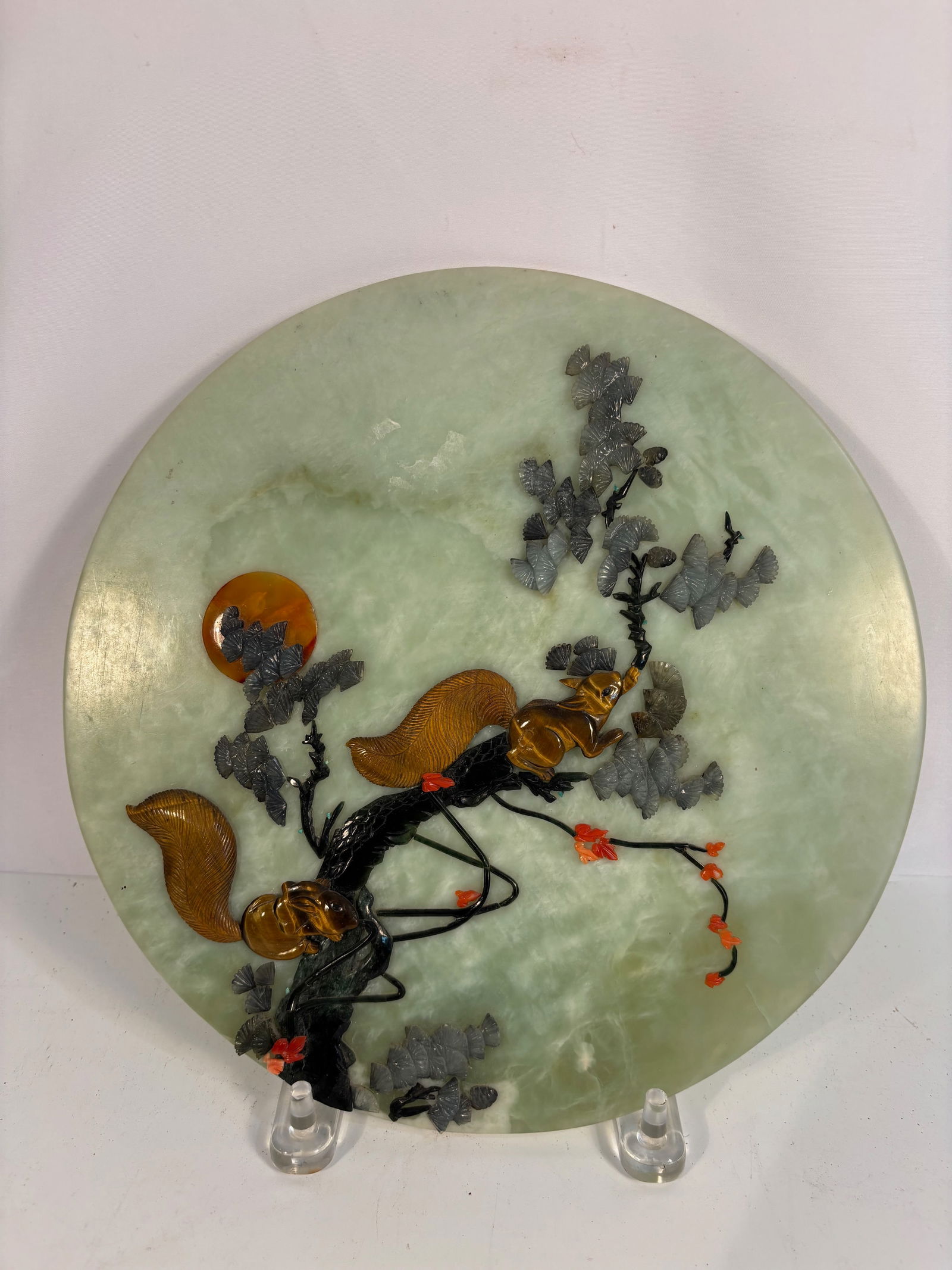 A Chinese Jade Plaque Inlaid with Coral, Agate, and Tiger’s Eye: A circular plaque of pale celadon jade, finely inlaid with coral, agate, and tiger’s eye stones to depict a lively squirrel climbing among grapevines with clusters of fruit. The squirrel’s body is