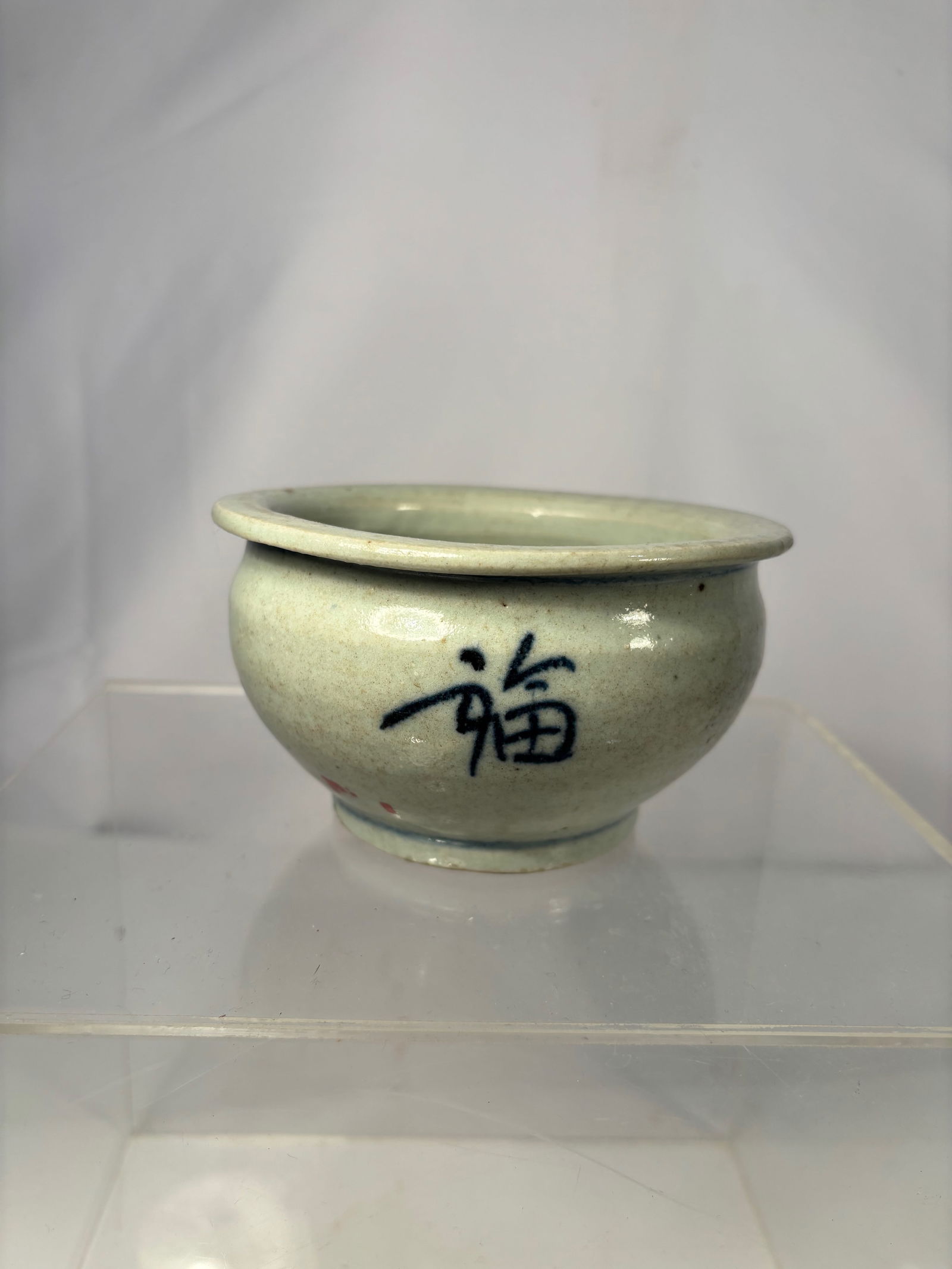 A Chinese Celadon-Glazed Bowl with Shou Character, 19th century: A Chinese celadon-glazed bowl of rounded form, the exterior painted in underglaze blue with a single Shou (longevity) character. Covered in a pale green celadon glaze with visible kiln texture and nat