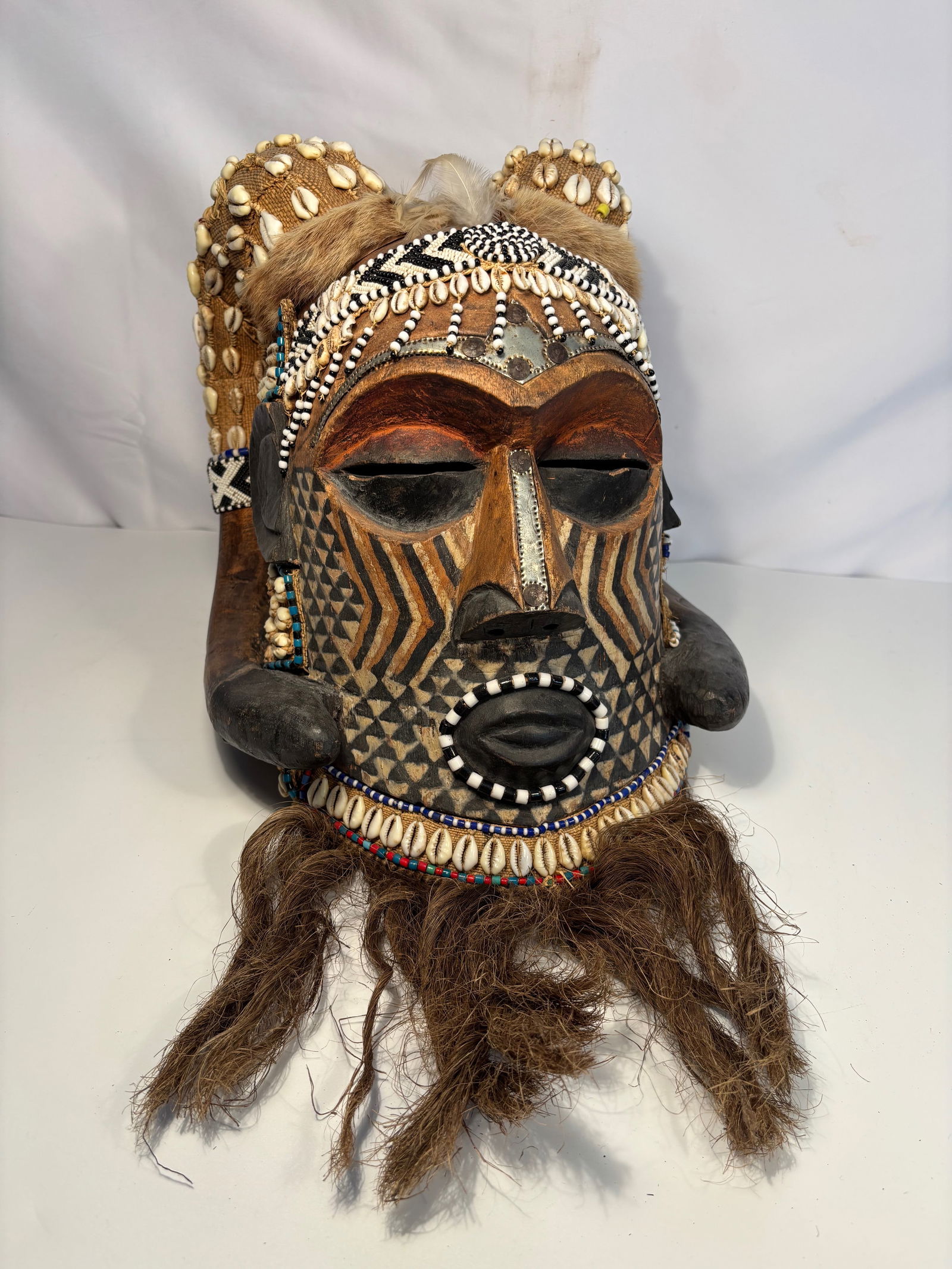 A Kuba Beaded Cowrie Shell and Fur Helmet Mask, Democratic Republic of Congo: A striking Kuba beaded helmet mask from the Democratic Republic of Congo, richly decorated with cowrie shells, glass beads, natural fibers, and animal fur. The carved wooden face exhibits bold geometr