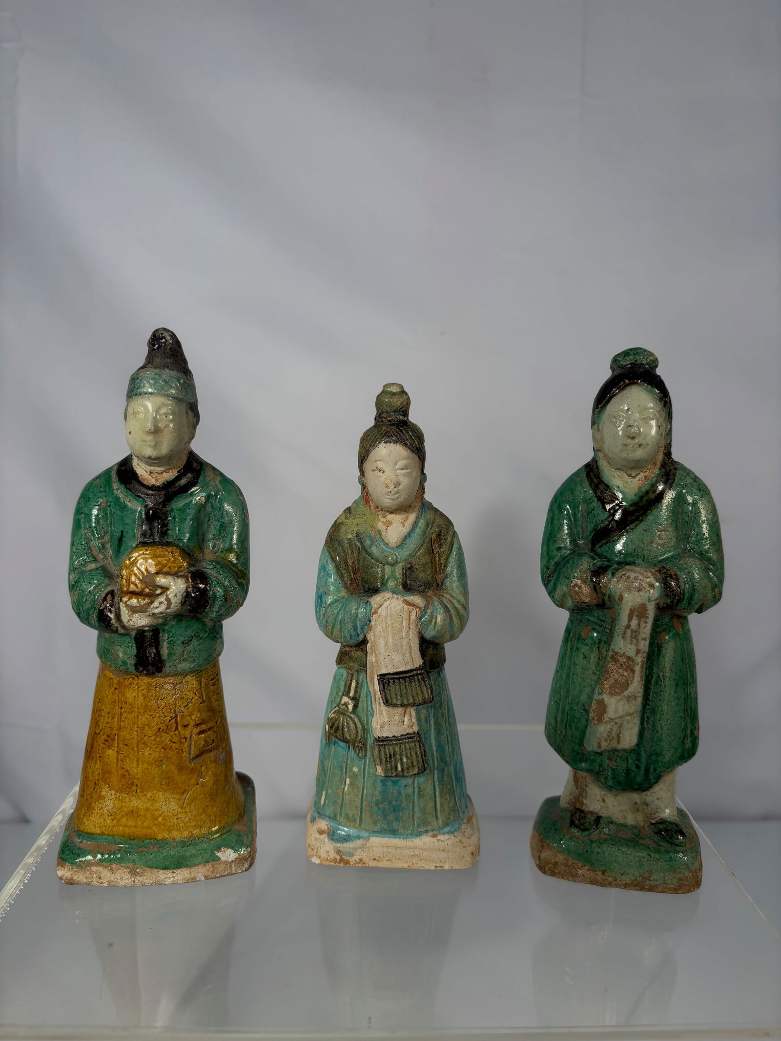 Set of Three Chinese Sancai-Glazed Pottery Attendant Figures, Ming Dynasty: Each figure modeled standing in long robes, glazed in green, amber, and cream tones. One holds an offering, the others with hands clasped in front. Flat bases with firing vents beneath. These tomb fig