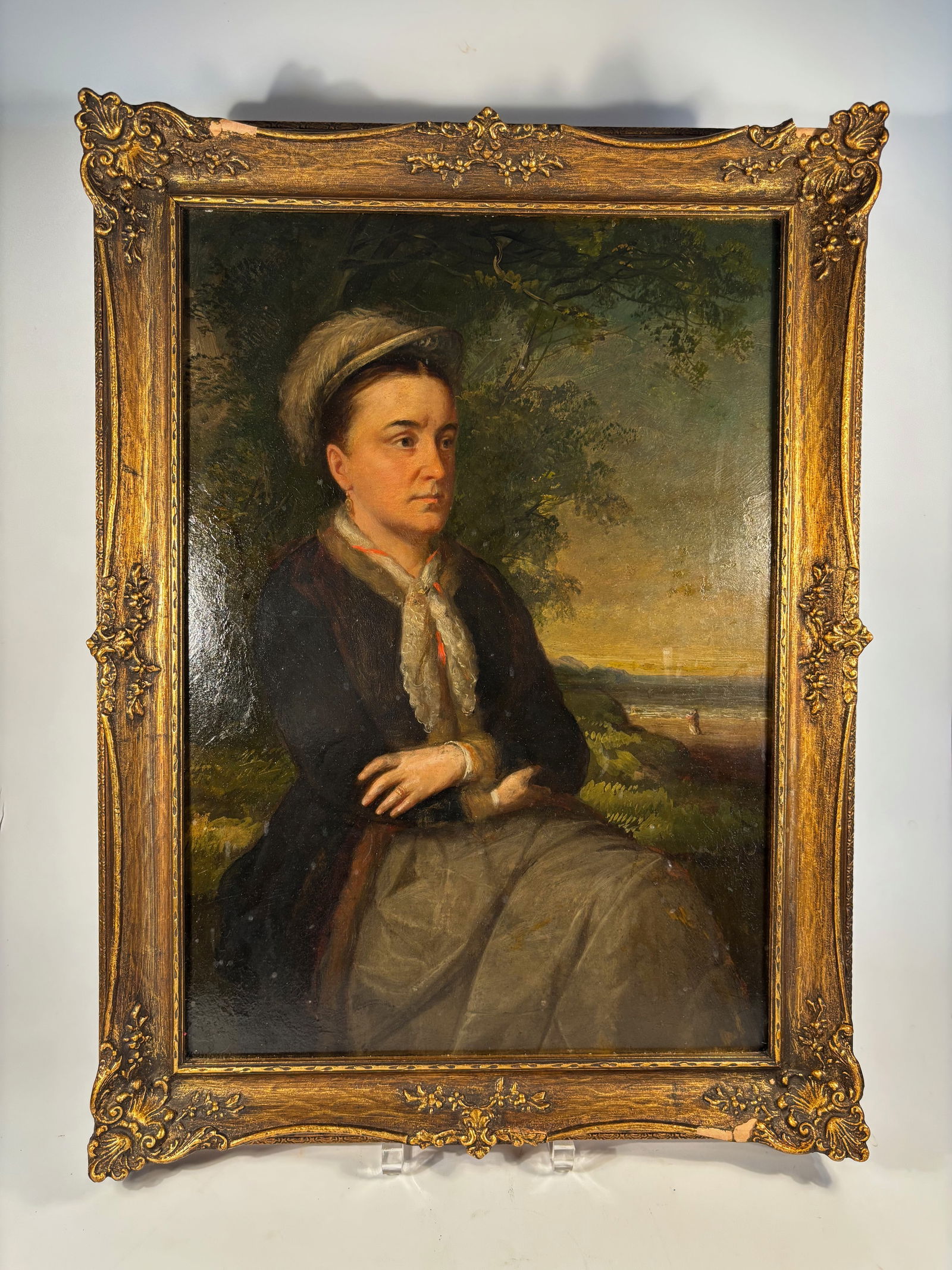 An English School Oil Portrait of a Seated Woman in Landscape, in Ornate Gilt Frame: 19th Century, portrait depicting a seated woman in a dark dress with lace scarf and bonnet, set against a soft wooded landscape with distant shoreline. The sitter’s calm expression and detailed atti