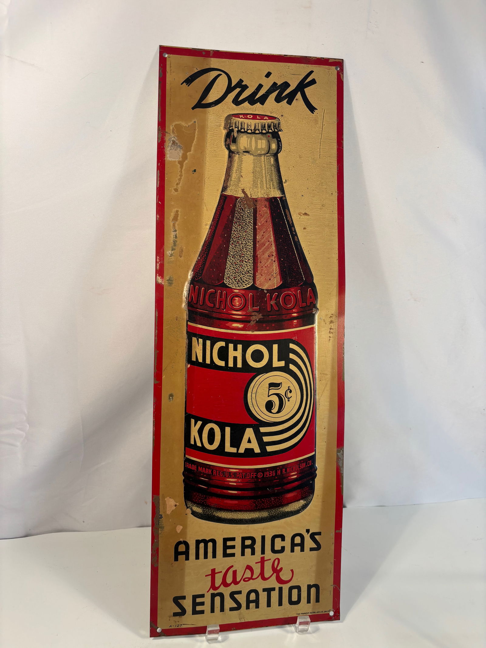 A 1930s Nichol Kola 5¢ Embossed Tin Lithograph Advertising Sign – “America’s Taste: Original Nichol Kola embossed tin lithograph advertising sign, circa 1936–1940, featuring the classic “Drink Nichol Kola 5¢ – America’s Taste Sensation” slogan. Made of tin-plated