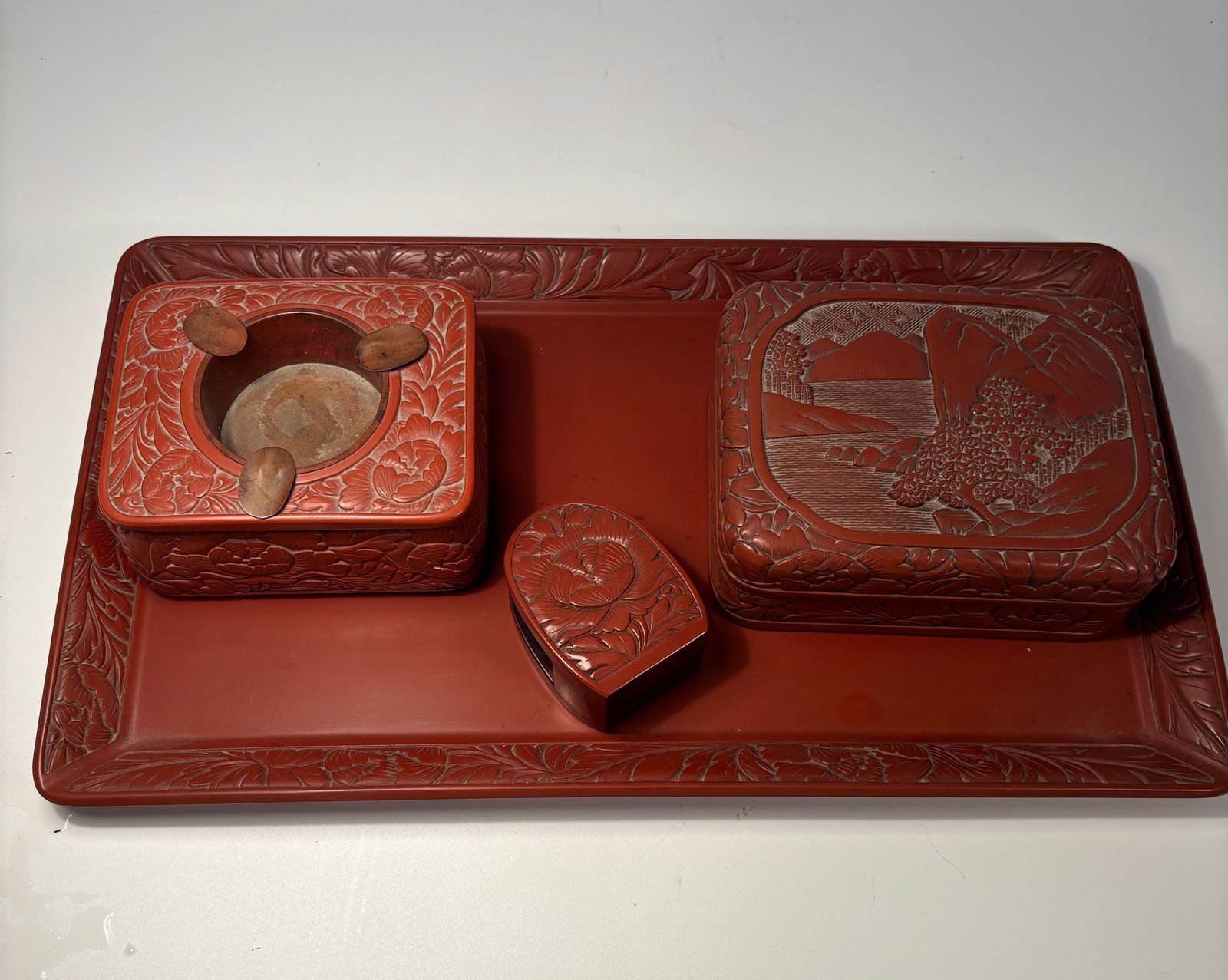 A Four-Piece of Japanese Red Cinnabar Tsuishu Lacquer Writing Set: Meiji period(1868-1912) , Comprising a rectangular tray, a lidded stationery box finely carved with a lakeside mountain landscape, a square inkstone box, and a smaller lidded box, each decorated in re