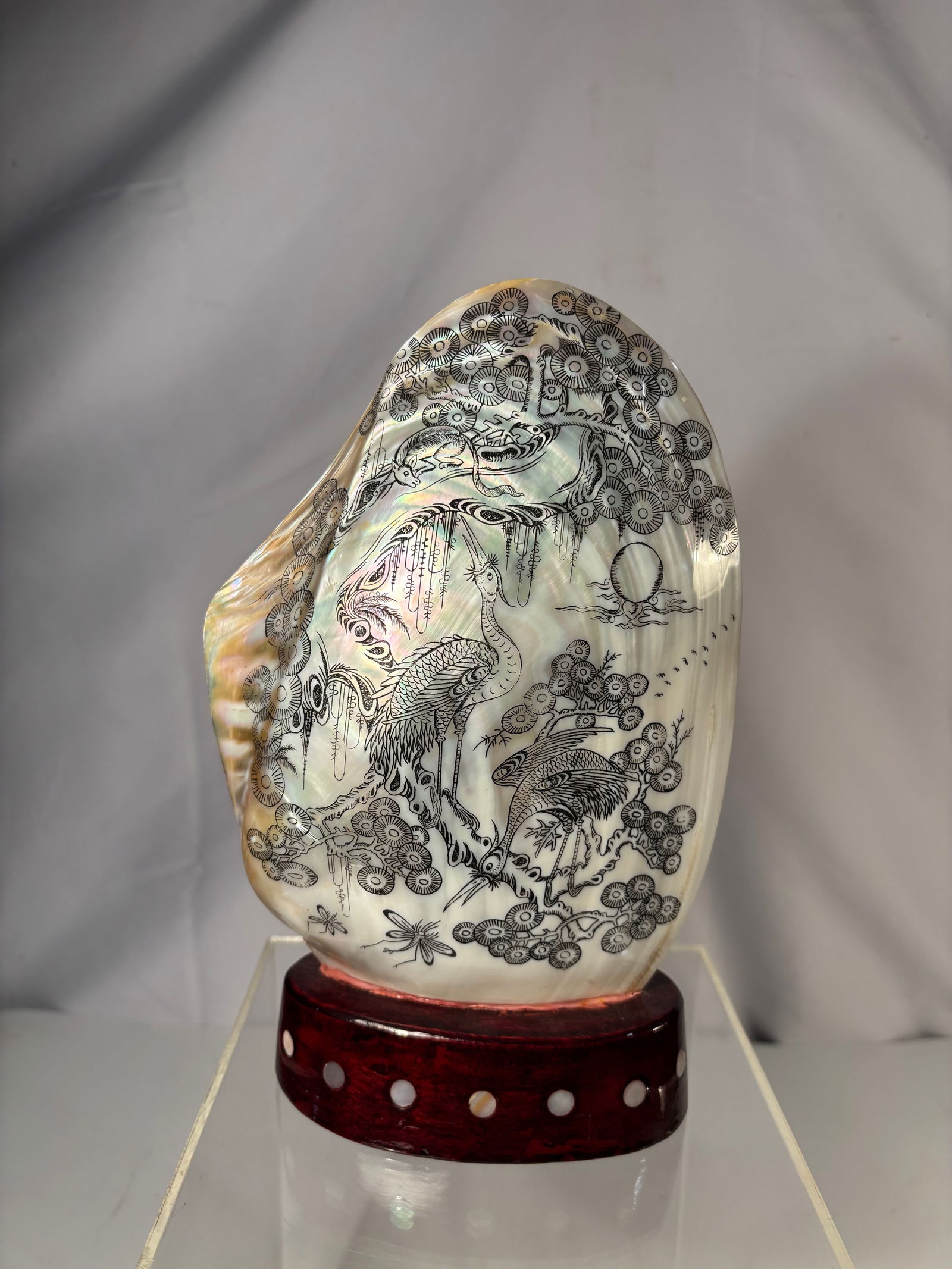 A Chinese Engraved and Painted Mother-of-Pearl Shell with Crane and Pine Design on Stand: Decorated mother-of-pearl shell featuring finely engraved and painted imagery of cranes among pine trees. The iridescent surface of the shell enhances the detailed linework and gives depth to the land
