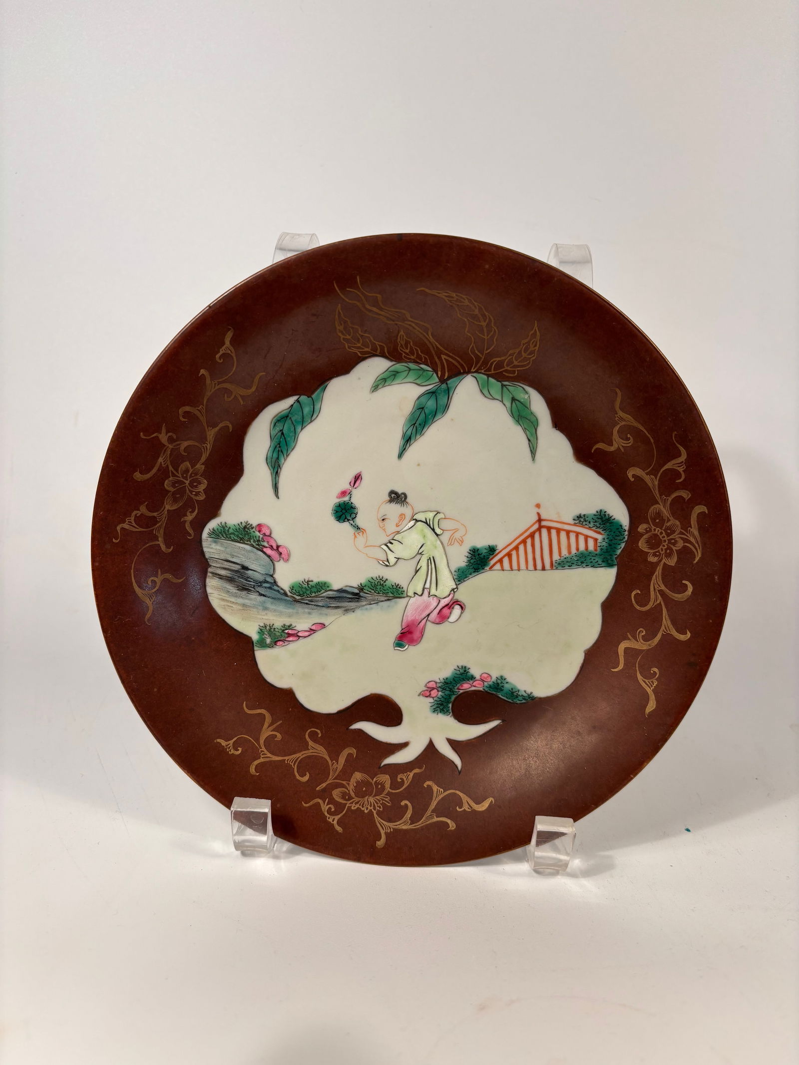 A Chinese famille rose porcelain plate with "Qianlong Mark": Qianlong period (1736-1795); A child in a garden setting holding a flower, surrounded by a brown-ground enamel border with gilt floral scrollwork — a theme symbolic of innocence, prosperity, and