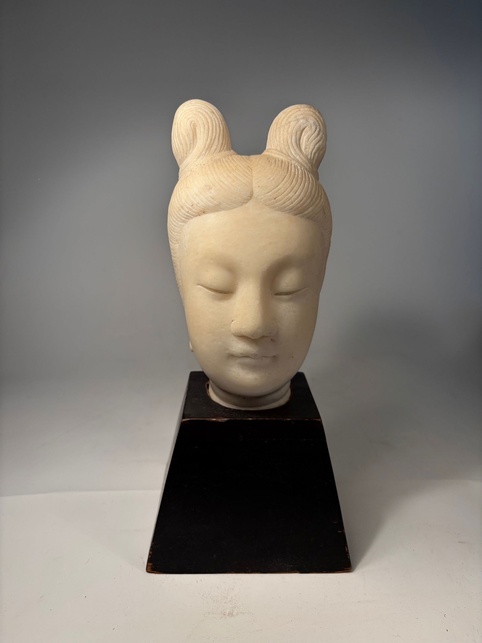 A Carved Stone Head of a Tang Dynasty-Style Lady on Stand: Tang dynasty (618–907 AD) or later; A carved stone head of a Tang-style lady, featuring a tranquil expression and high twin-bun coiffure. The stone displays natural patina and weathering on the reve