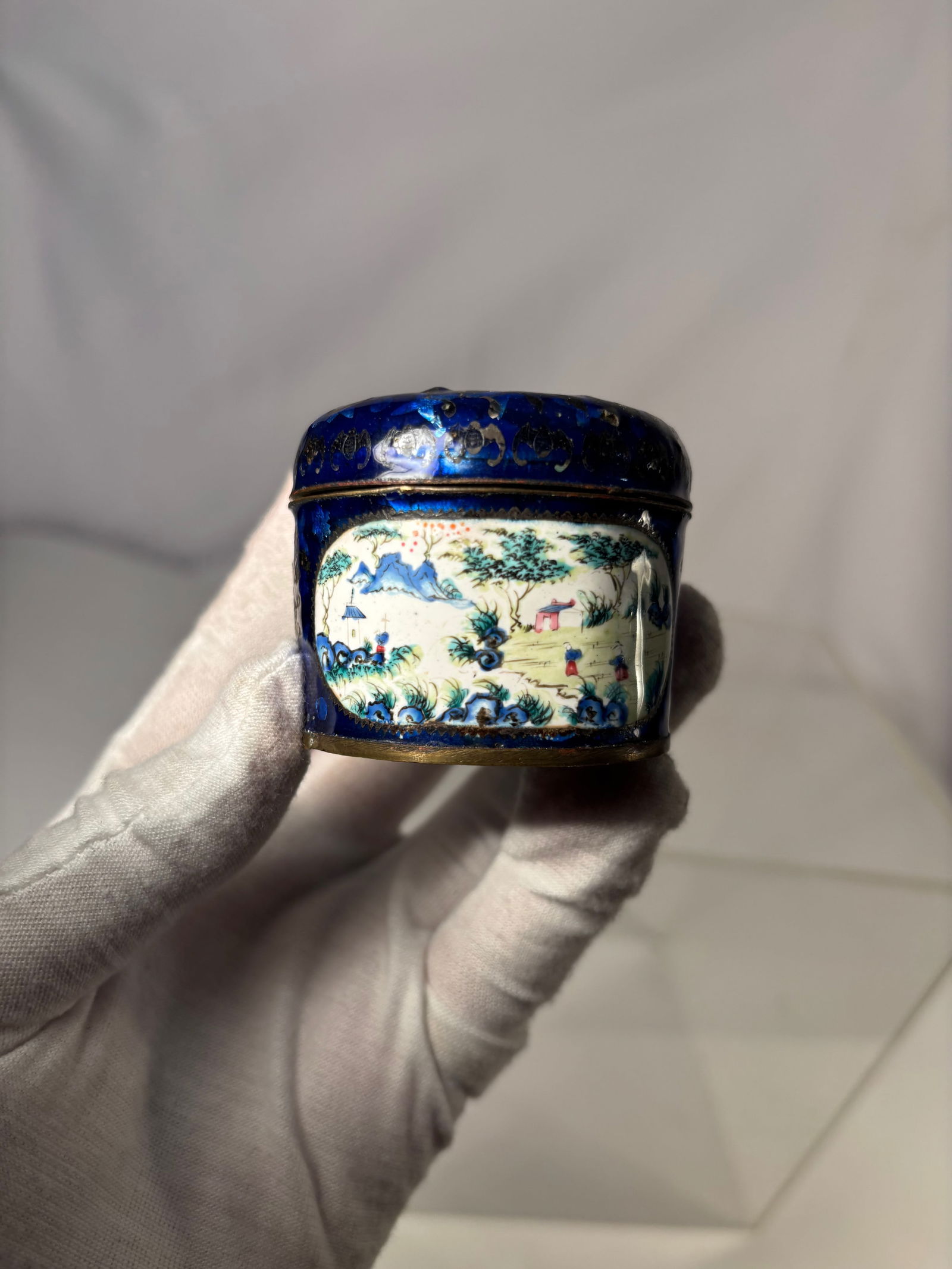 A Chinese Blue Enamel Lidded Snuff Box with Figural Landscape: Qing dynasty(1644-1912); A Chinese enameled metal snuff box of cylindrical form, decorated on the exterior in deep cobalt-blue enamel and centered with a painted famille rose–style landscape panel d
