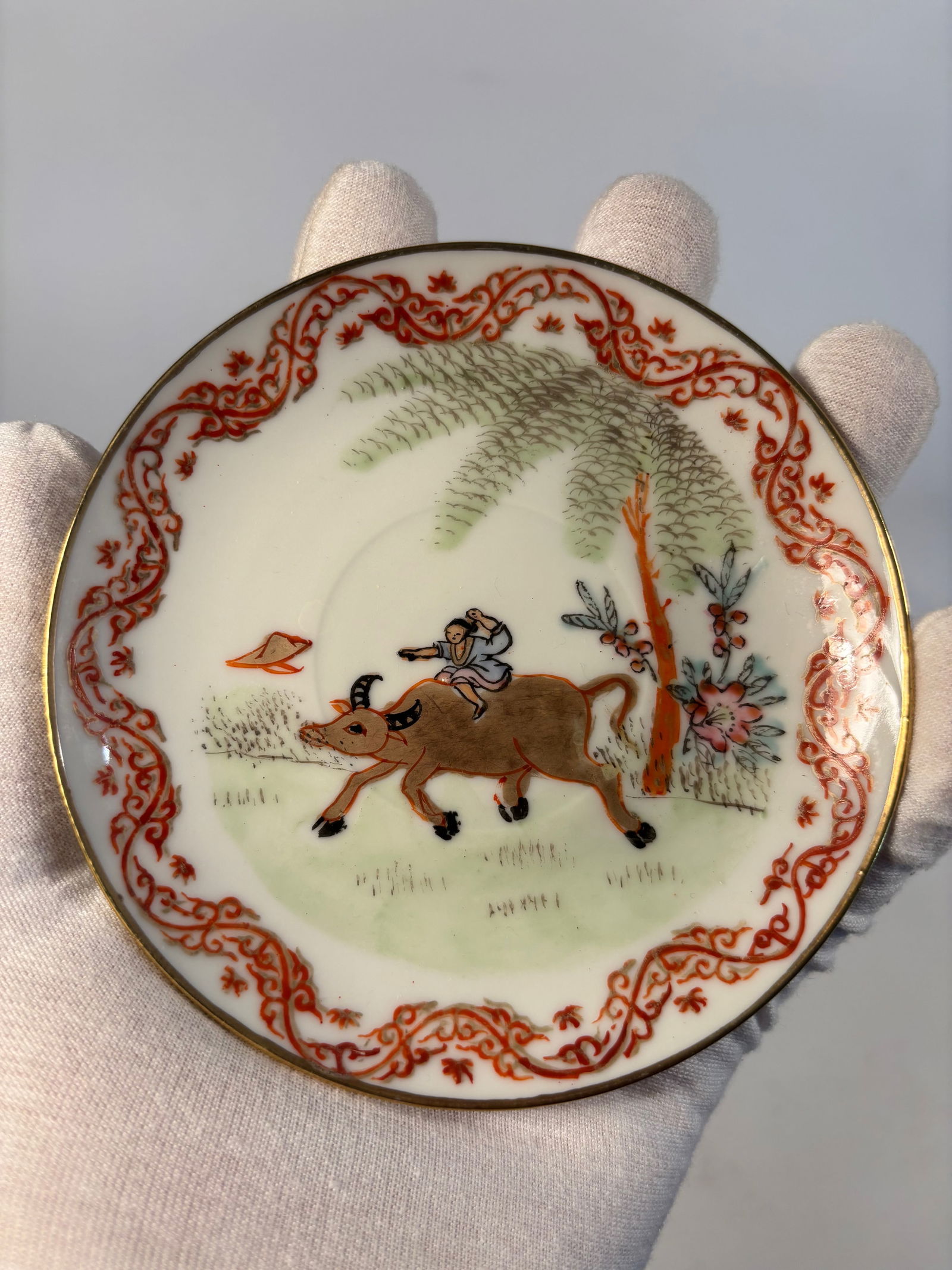 A Hand-Painted Porcelain Saucer with Boy on Water Buffalo: Late 19th century -early 20th century,The central scene depicts a boy riding a water buffalo beneath a large willow or palm tree, a classic Chinese pastoral motif symbolizing tranquility and harmony