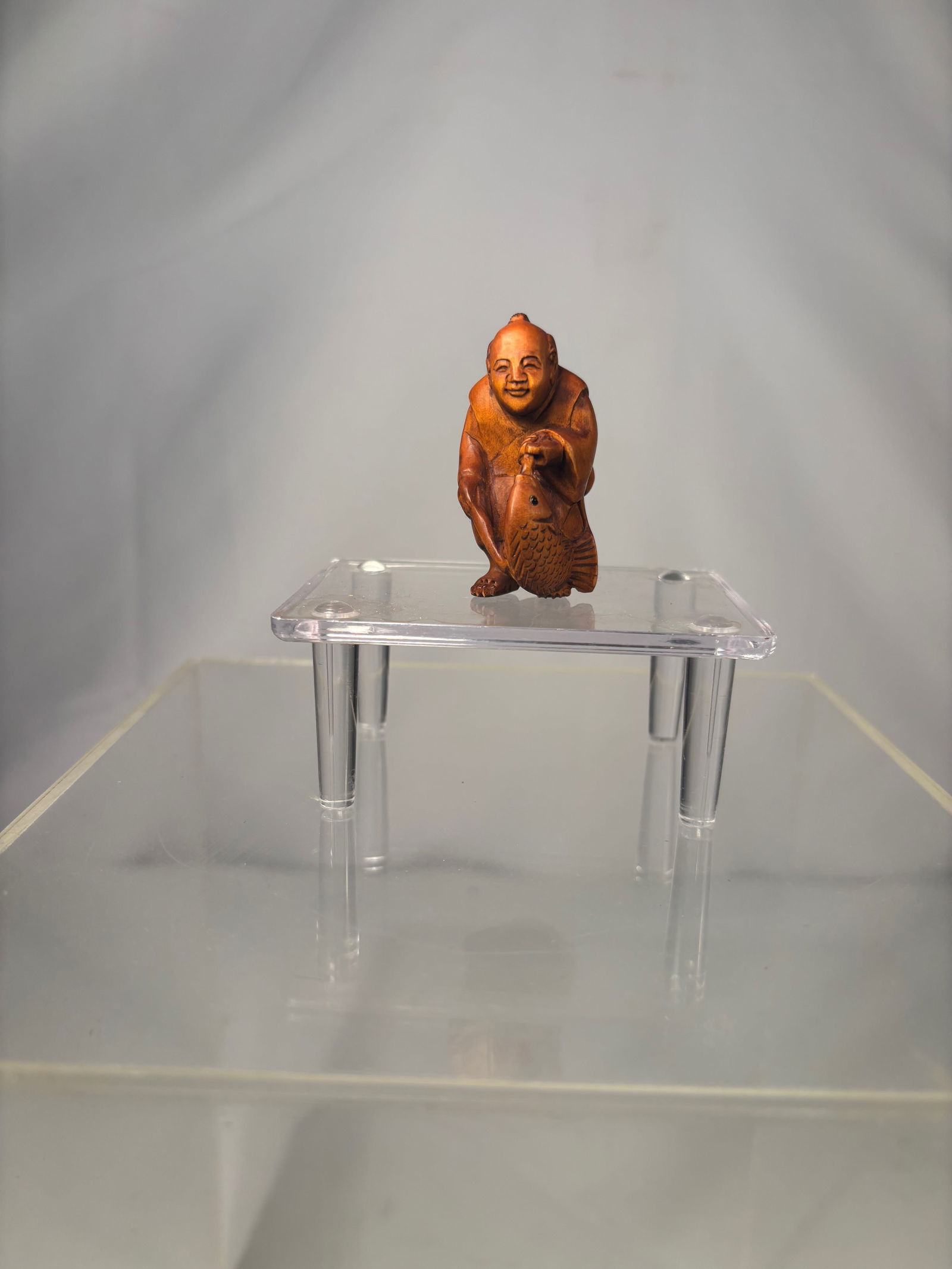 A Japanese Boxwood Netsuke – Smiling Itinerant Monk Carrying a Fish, Matsuyama Mark: Meiji period (19th century), Finely carved Japanese boxwood netsuke depicting a smiling itinerant monk standing upright and carrying a large fish over his shoulder. The figure is rendered with express