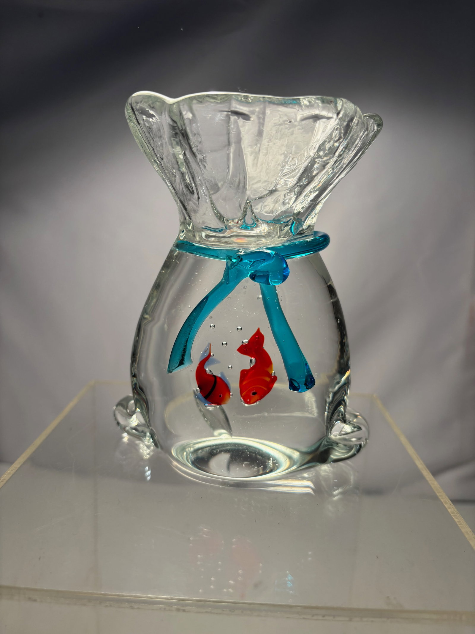 An Art Glass Drawstring Bag-Form Vase with Encased Fish, Late 20th Century: Decorative art glass vase formed in the shape of a drawstring bag, executed in clear glass with applied blue glass tie decoration at the neck. The interior encloses two small red glass fish, suspended