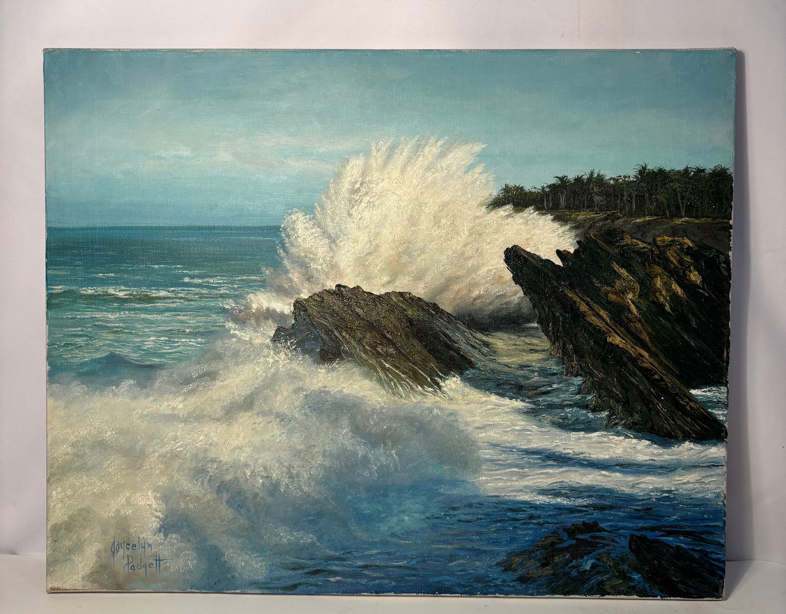 A Signed Coastal Seascape Painting with Waves Crashing on Rocks, Joycelyn Padgett: Original coastal seascape painting depicting waves crashing against a rocky shoreline beneath a blue sky. Executed with energetic brushwork and visible surface texture. Signed lower right “Joycelyn