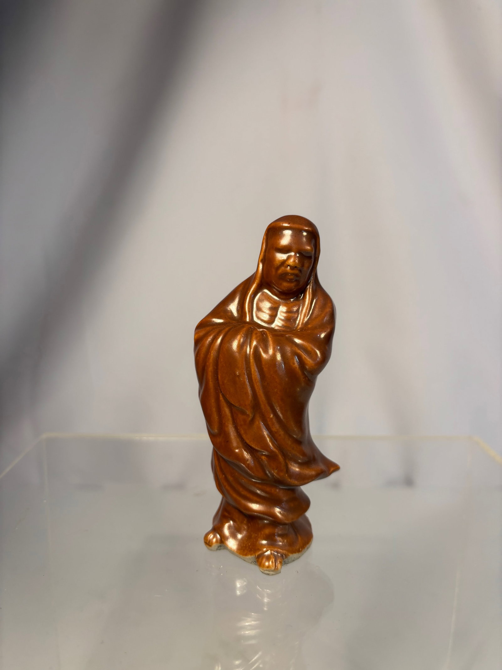 A Chinese Brown-Glazed Ceramic Figure of Bodhidharma: Qing dyansty(1644-1912); Well modeled in flowing robes with a serene expression, the figure stands with arms folded beneath his draped mantle. The rich brown glaze has a soft lustrous sheen, pooling i