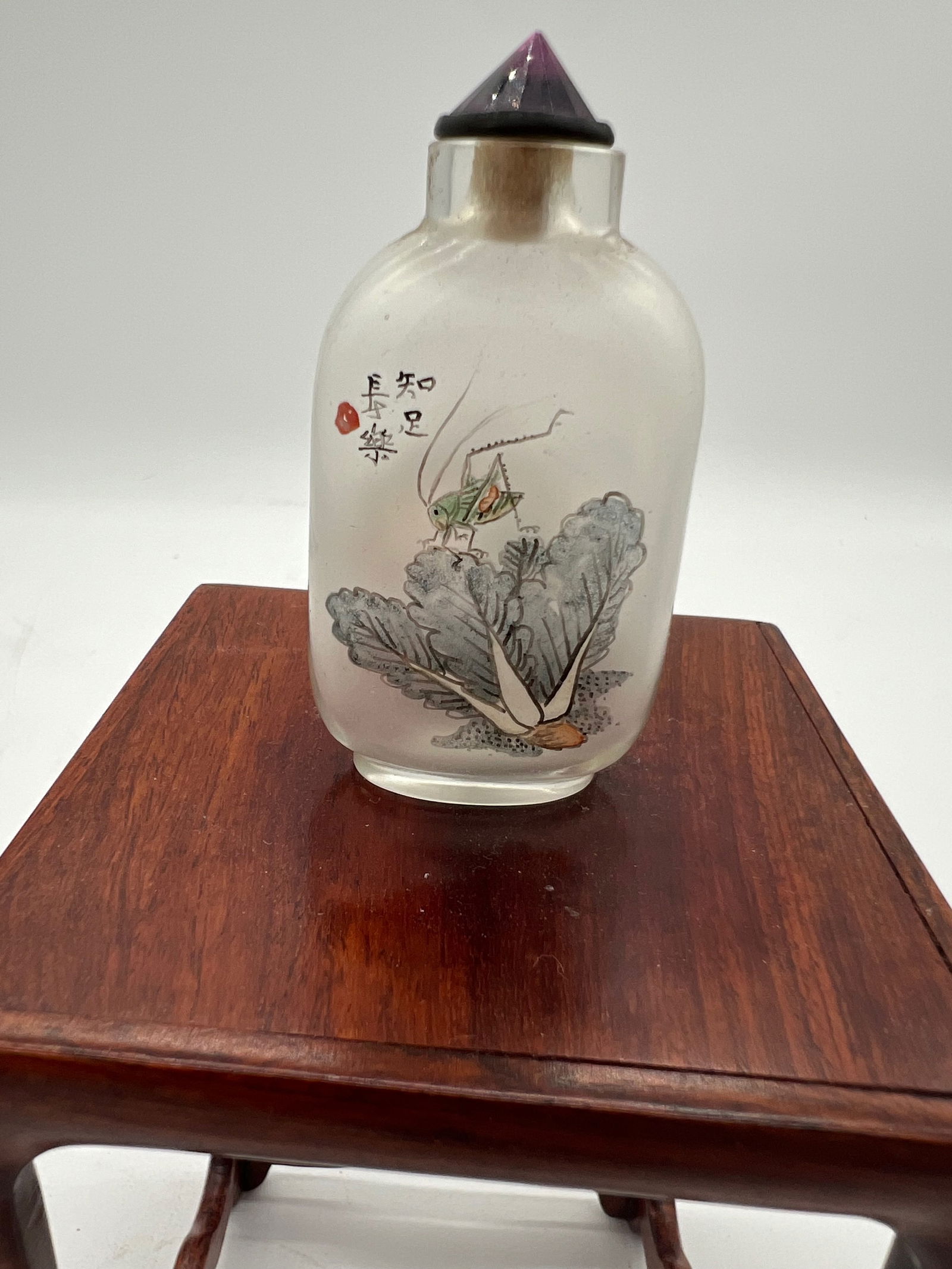 A Chinese inside-painted glass snuff bottle: Qing Dynasty (1644–1912) Side 1: Depicts a grasshopper on bok choy, symbolizing simplicity, humility, and good fortune in Chinese culture. Side 2: A traditional landscape scene, with a tree, house,