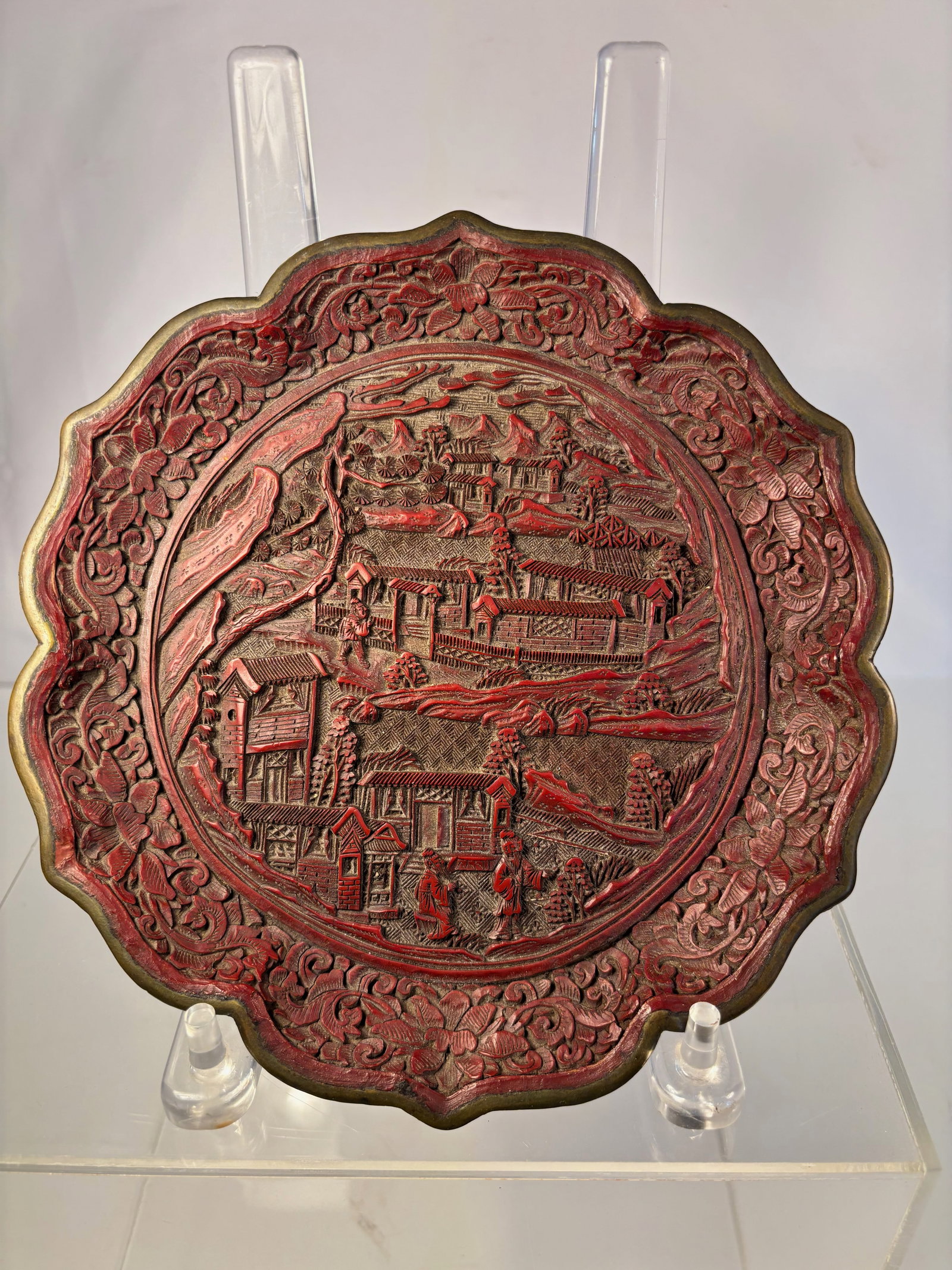 A Chinese Carved Cinnabar Lacquer Dish with Landscape and Figures: Qing dynasty(1644-1912); A finely carved Chinese cinnabar lacquer dish of lobed form, decorated in high relief with a continuous landscape scene of pavilions, bridges, and figures among pine trees and