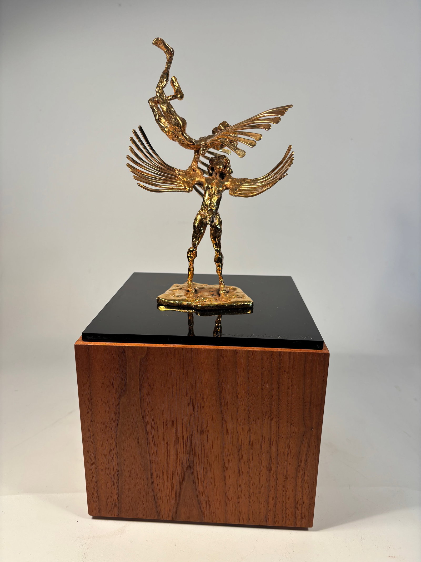 Joseph A. Burlini (American, b. 1937) Gilded Bronze Winged Figures Sculpture, signed and dated 1982: A striking bronze composition featuring a stylized winged male figure hoisting an acrobatic nude form overhead—symbolizing flight or transcendence—mounted on a high-gloss black acrylic plinth. Inc
