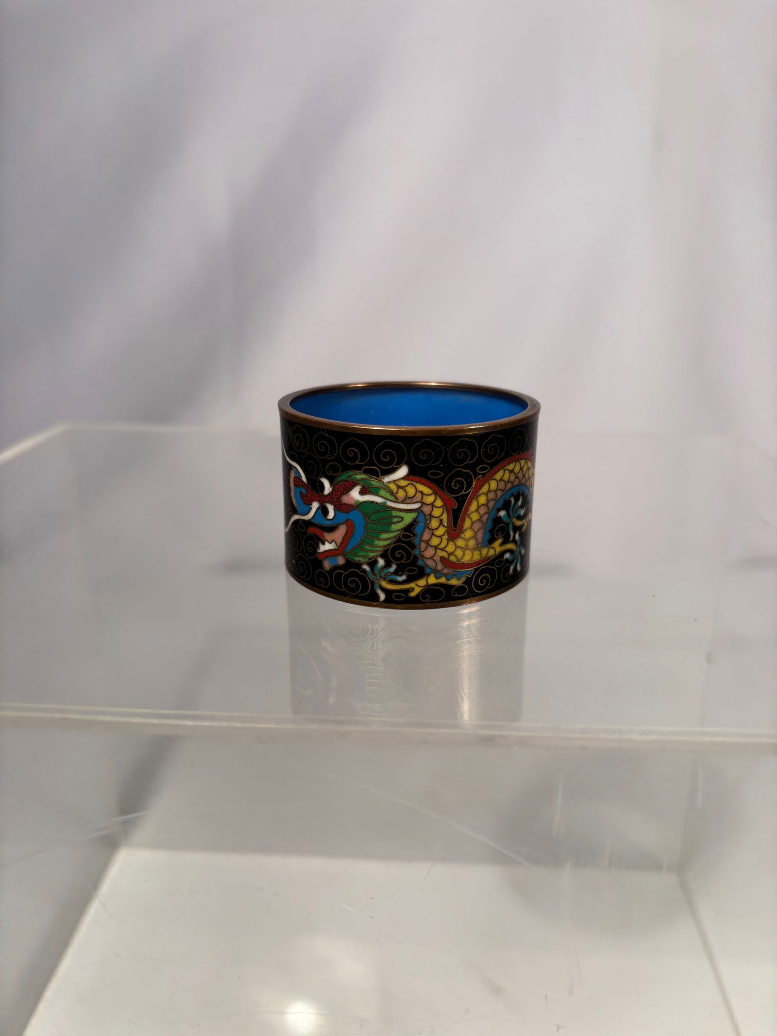 A Chinese Cloisonné Dragon Motif Napkin Ring with Blue Interior: Qing Dynasty (1644-1922); Chinese cloisonné enamel napkin ring of cylindrical form, decorated with a colorful dragon amid scrolling cloud motifs on a black ground. The design is outlined with fine me