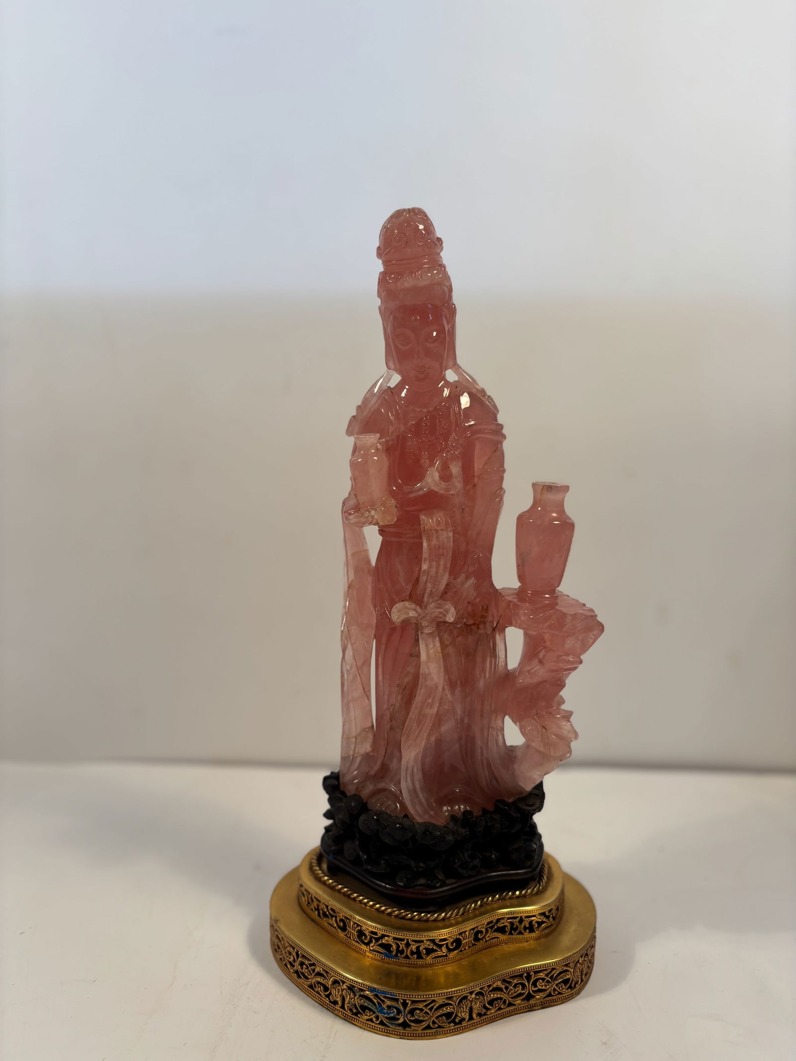 A Chinese carved rose quartz figure of Guanyin Statue with base: Late 19th century to early 20th century; The figure depicts Guanyin (the Bodhisattva of Compassion) standing in flowing robes, holding a vase and scroll, symbolizing mercy and wisdom. She stands on a