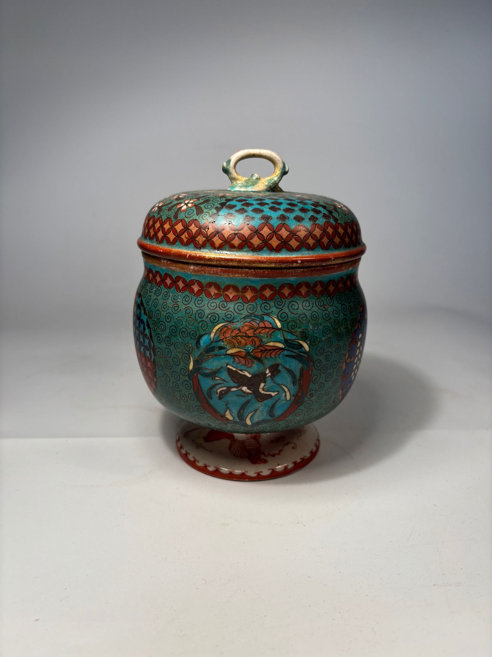 A Japanese Porcelain Lidded Jar with Cloisonné-Style Enamel Decoration: Early 20th Century; A vibrant Japanese lidded porcelain jar, decorated in a cloisonné-style enamel technique, featuring colorful floral and geometric designs in turquoise, rust-red, and black enamels