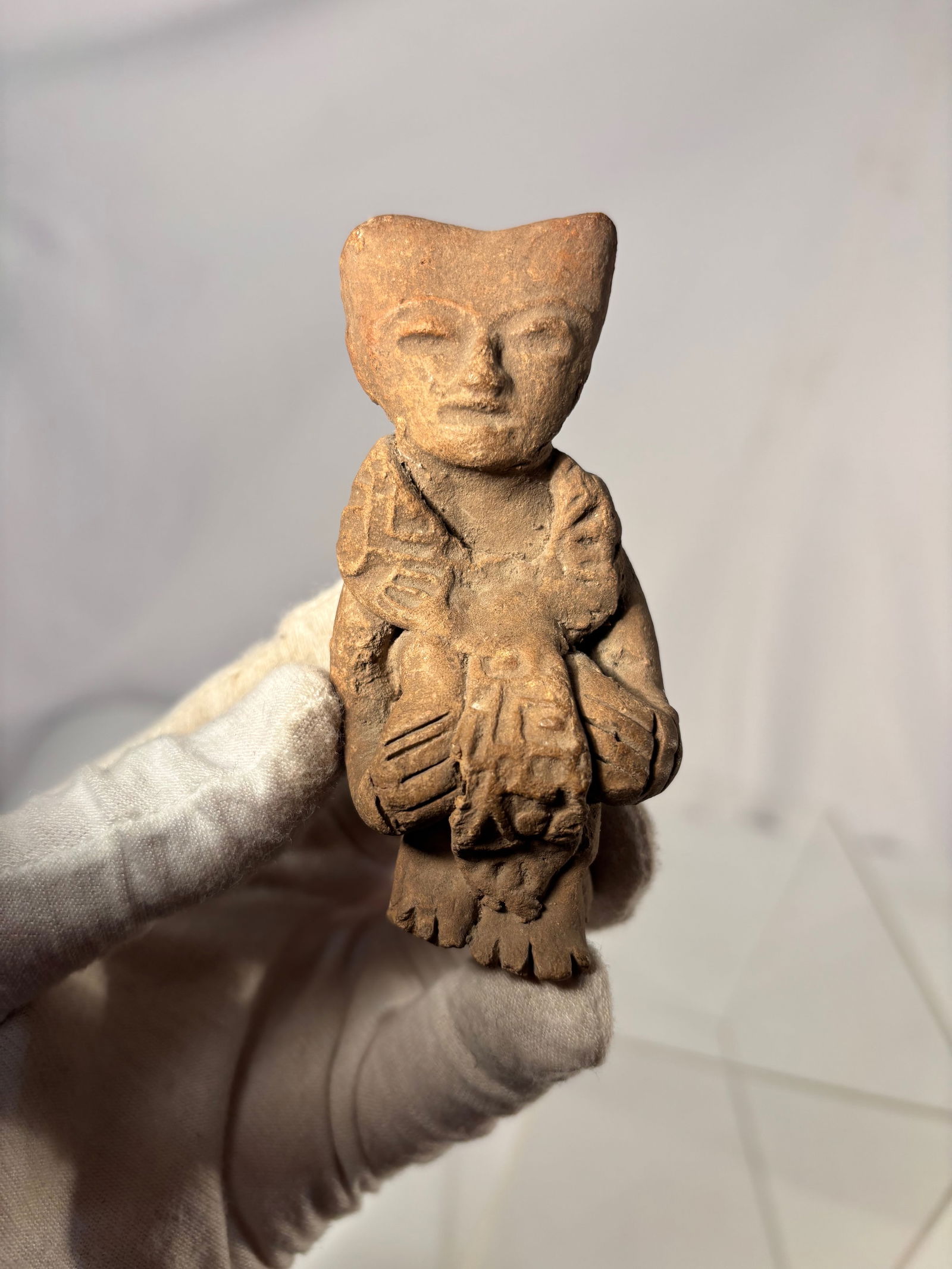 Pre-Columbian Terracotta Seated Figure, West Mexico: Small hand-formed terracotta figure in the style of ancient West Mexican cultures (Nayarit / Jalisco / Colima). The figure depicts a seated person with stylized triangular head, incised facial feature