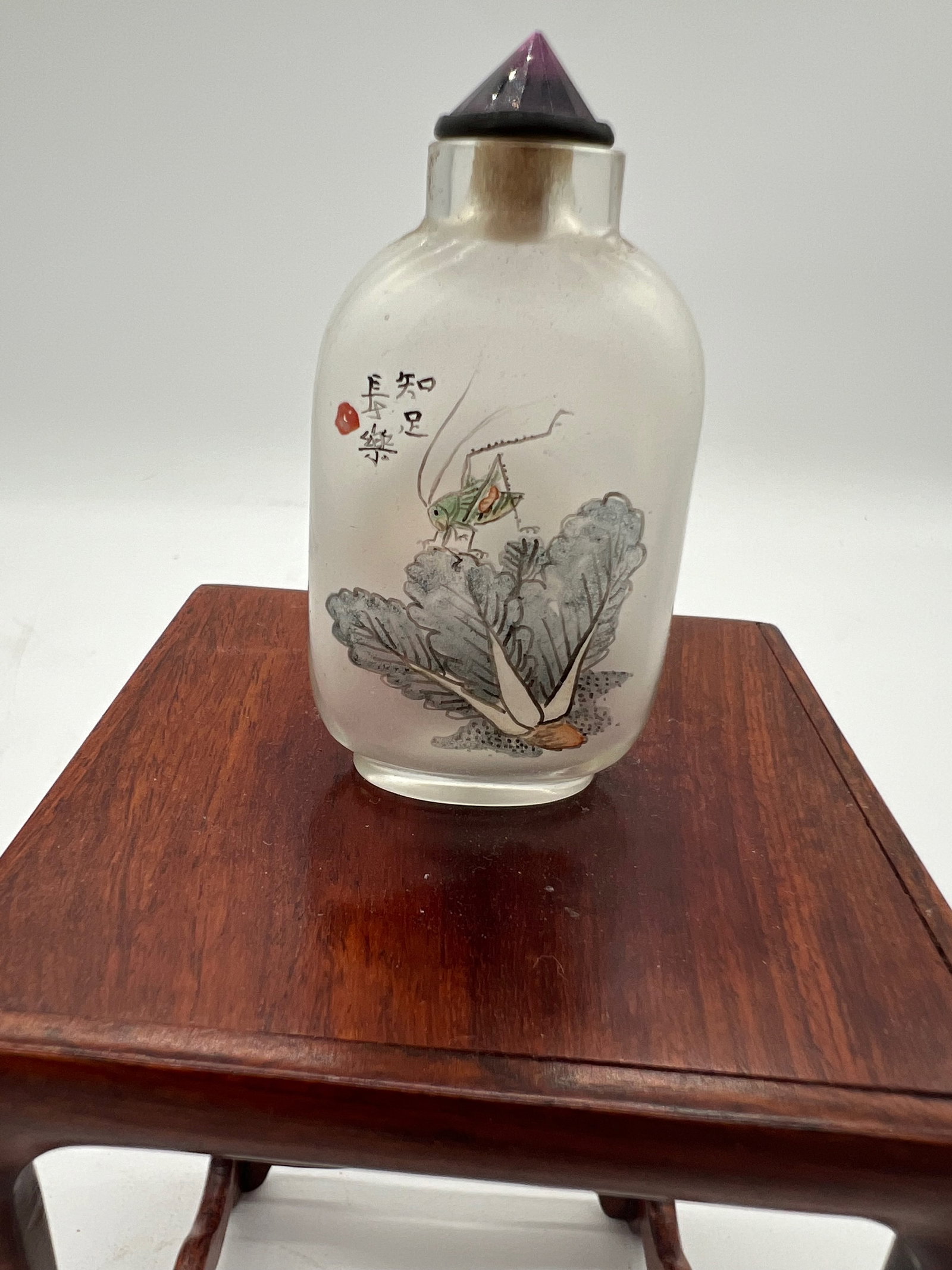 A Chinese inside-painted glass snuff bottle: Qing Dynasty (1644–1912) Side 1: Depicts a grasshopper on bok choy, symbolizing simplicity, humility, and good fortune in Chinese culture. Side 2: A traditional landscape scene, with a tree, house,