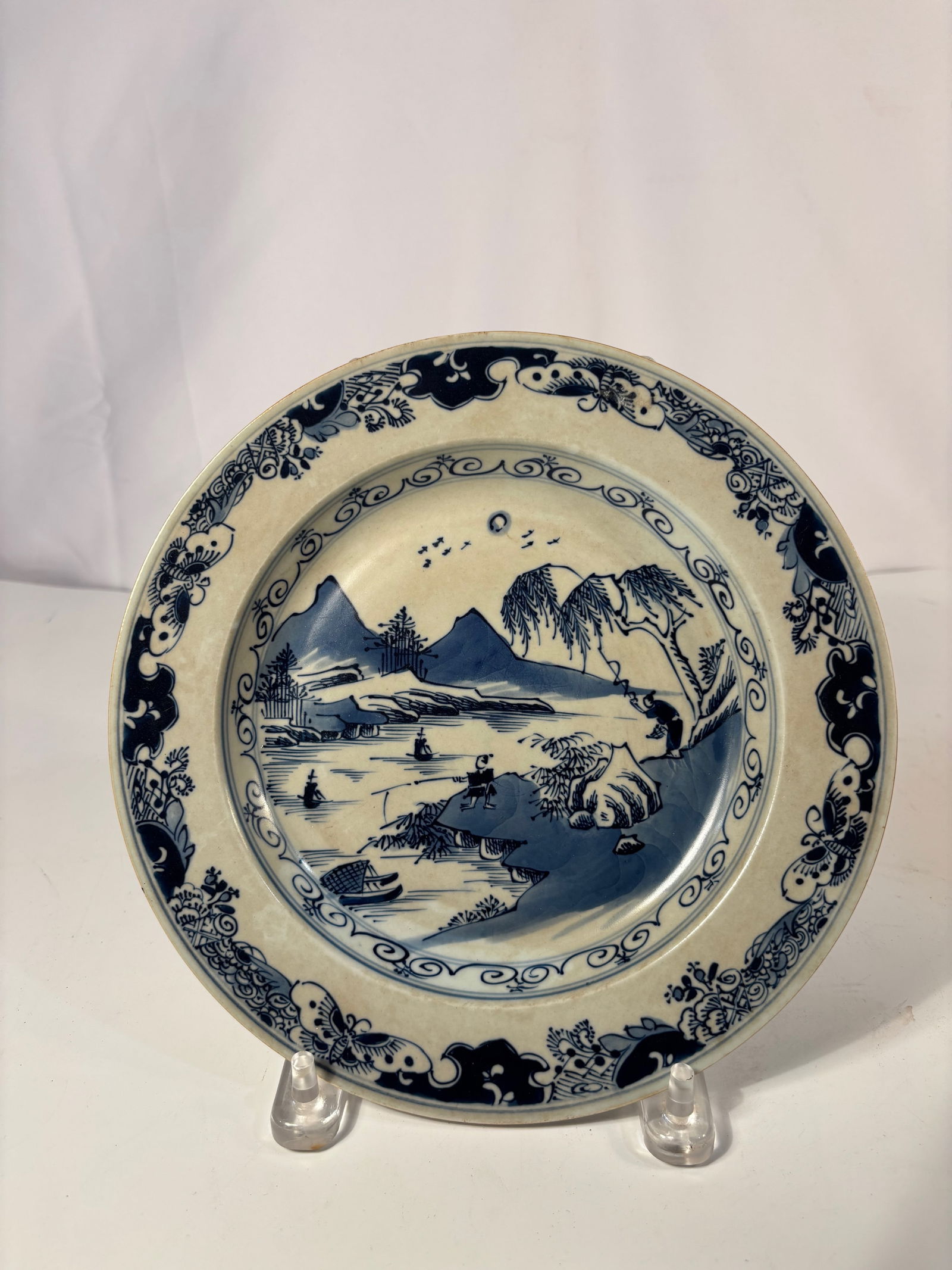 A Japanese Blue-and-White Porcelain Plate Meiji Period (c.1880–1910), Square Seal Mark: Finely painted Japanese blue-and-white porcelain plate depicting a river landscape with figures, pavilions, and sailing boats within a scrolling floral border. Glazed in soft white with deep cobalt