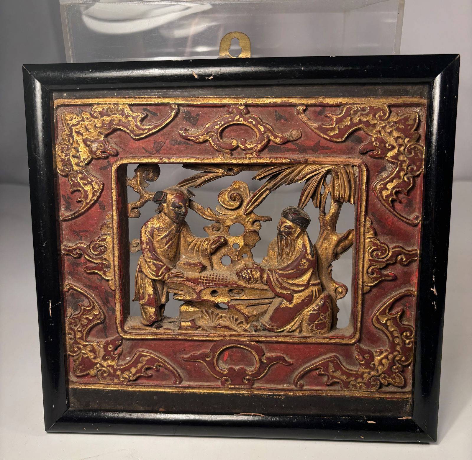 A Chinese Carved and Gilt Wooden Panel with Figures: Qing dynasty(1644-1912); A finely carved and polychrome-gilt wooden panel depicting two scholars or officials standing beside a table with scrolls and ritual vessels, set under a bamboo and pine canop