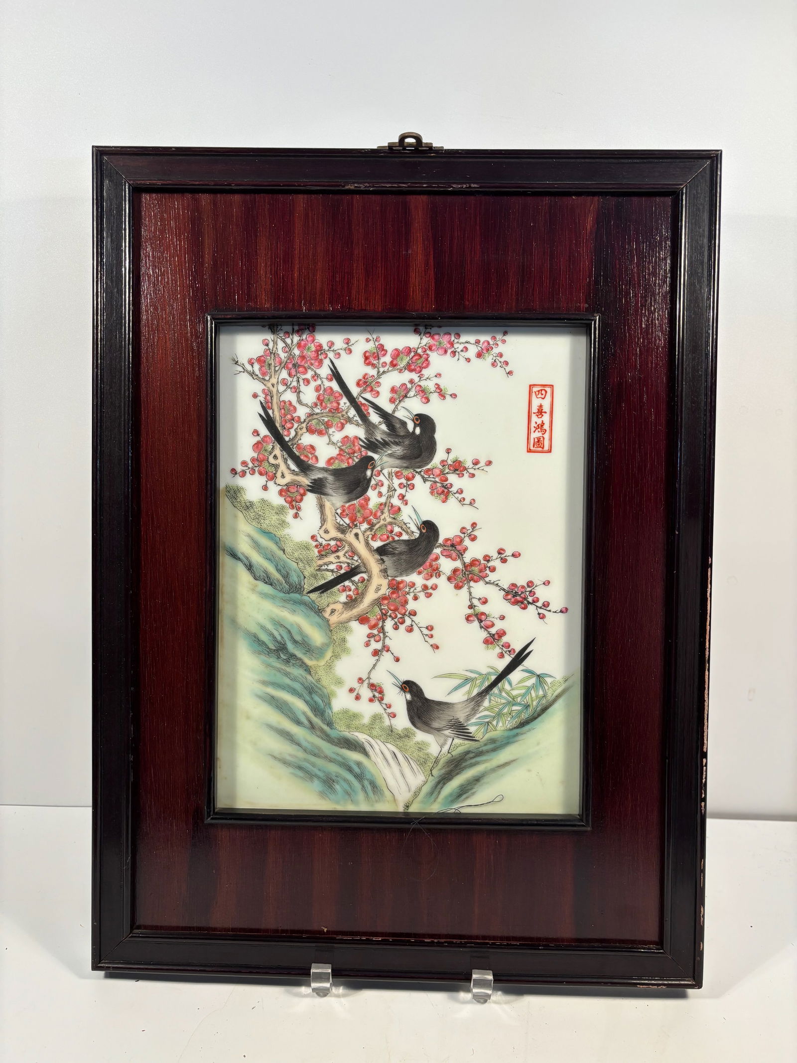 A framed Chinese hand painted porcelain plaque: Chinese porcelain plaque, framed and decorated in a traditional famille rose plaque. It features four birds perched among red plum blossoms. Dimensions?Frame width: 14.5 inches; Frame height: 19.5 inc