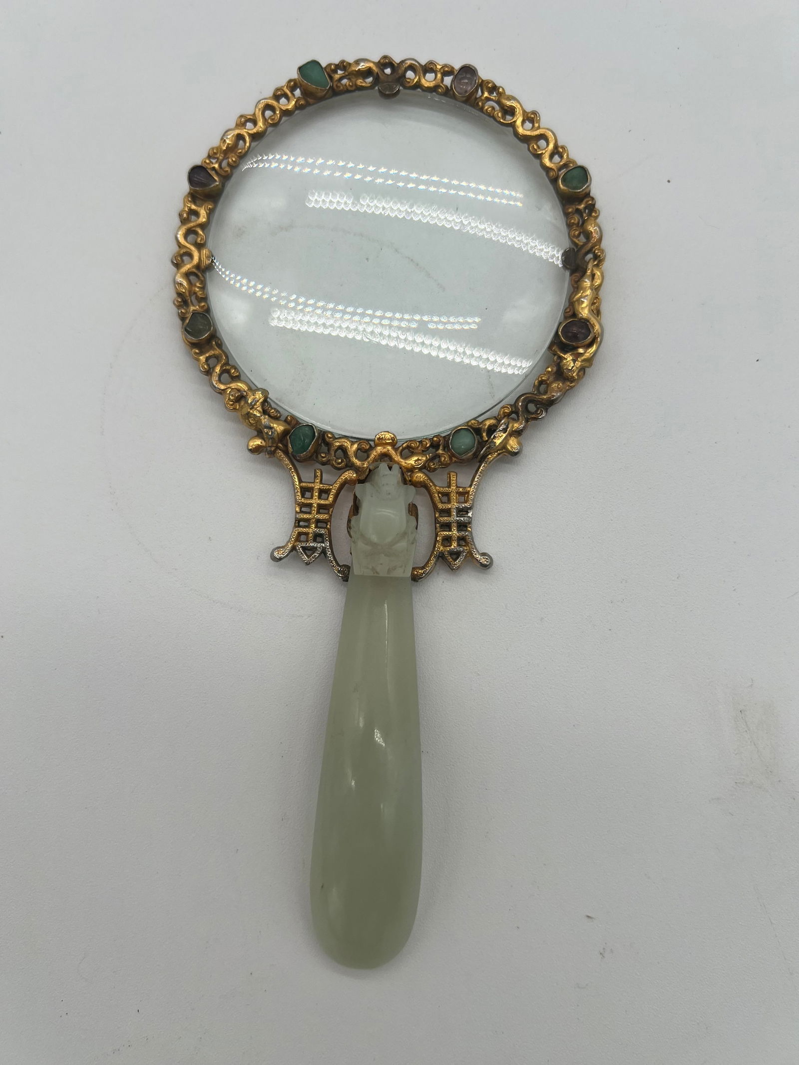 A gilt bronze framed magnifying glass with a jade handle and tourmaline gemstones: Qing Dynasty (1644-1912); The magnifying glass with a jade handle and is adorned 16 small gemstones like jadeite, tourmaline & etc. The frame surrounding the lens appears highly detailed, with a flora