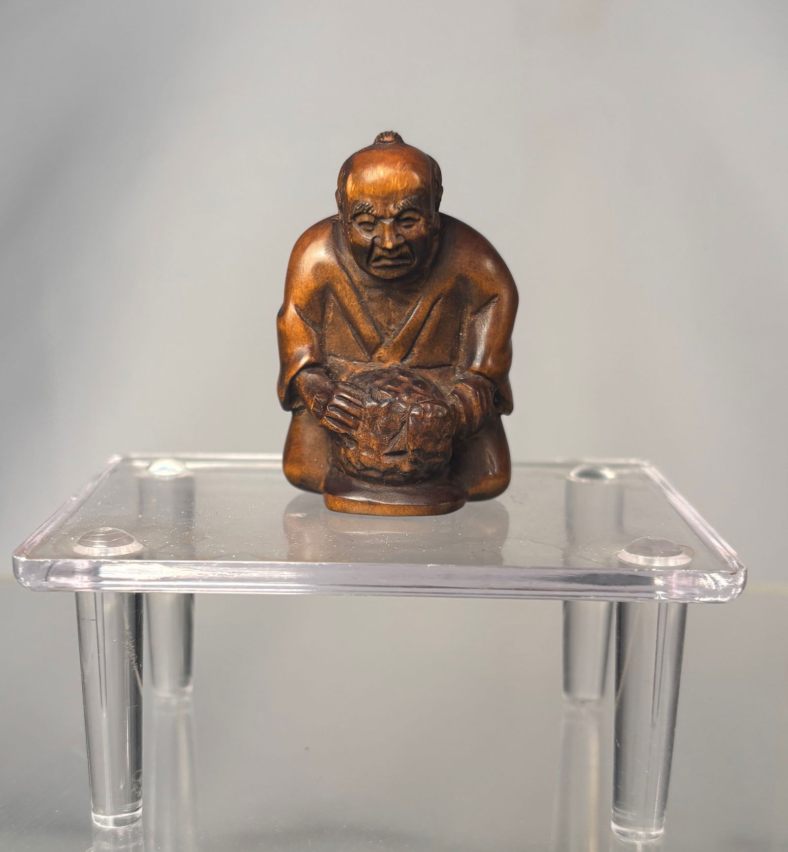 A Japanese Boxwood Netsuke of a Seated Oni on a Barrel, Signed (Gyokumatsu): Meiji period (19th century), A finely carved Japanese netsuke depicting a seated male figure, shown crouching with hands clasped before him, rendered in dense boxwood with a warm, well-developed patin