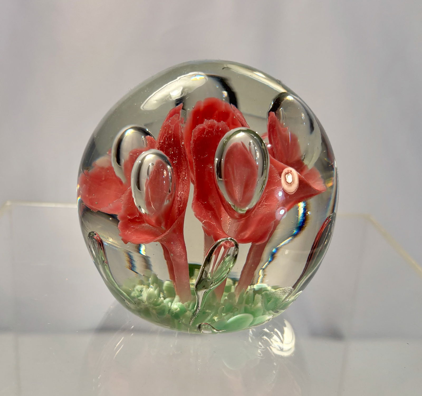 A Vintage Art Glass Paperweight with Red Flowers and Controlled Bubbles: A vintage hand-blown art glass paperweight featuring three vivid red lampwork flowers rising from a green ground. The clear glass dome contains multiple controlled air bubbles surrounding the floral s