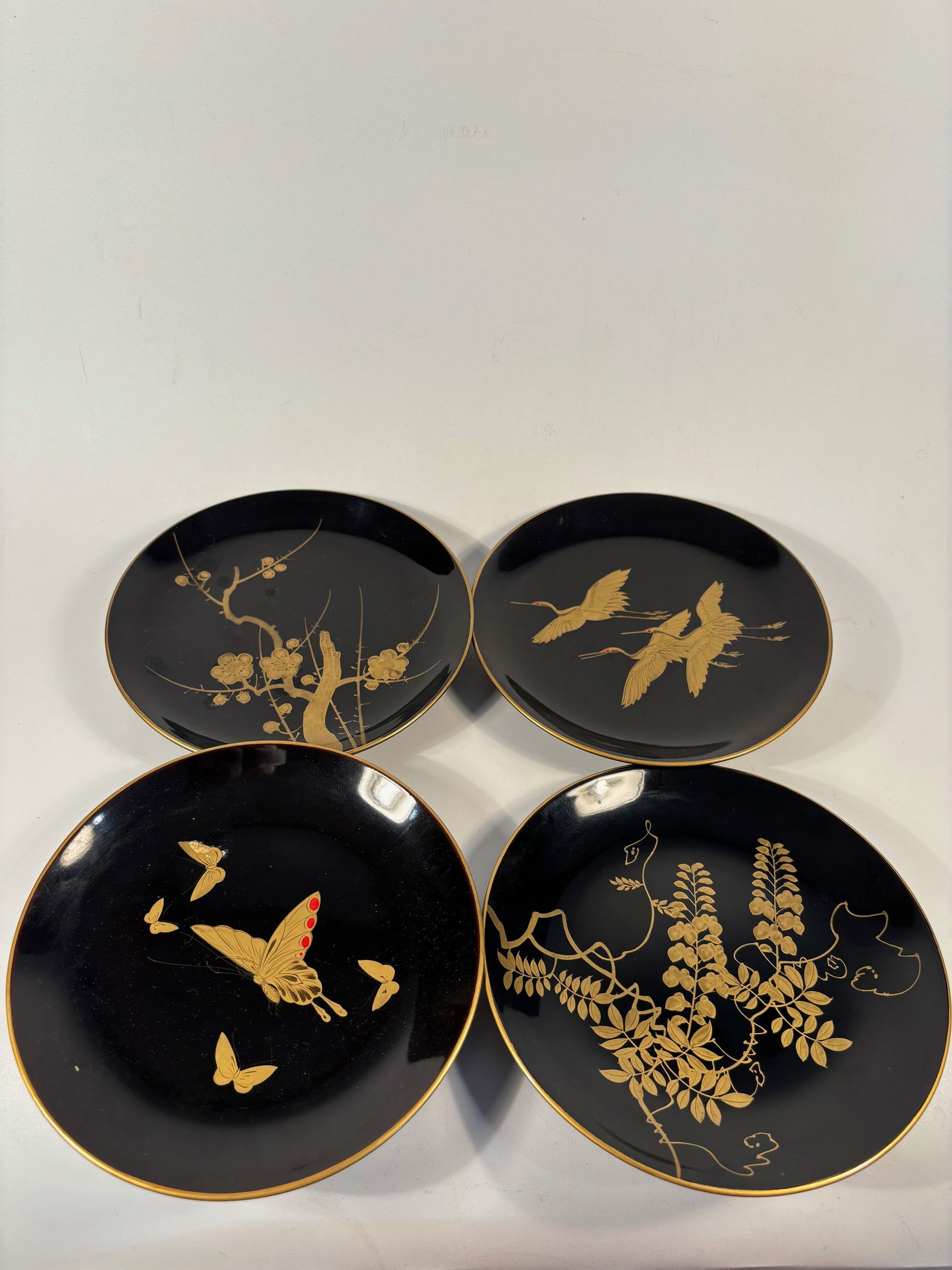 Set of 4 Japanese Makie Lacquer plates: Each plate finely decorated in gold maki-e on black lacquer with traditional Japanese motifs including plum blossoms, cranes in flight, butterflies, and flowering vines. The designs are delicately ren