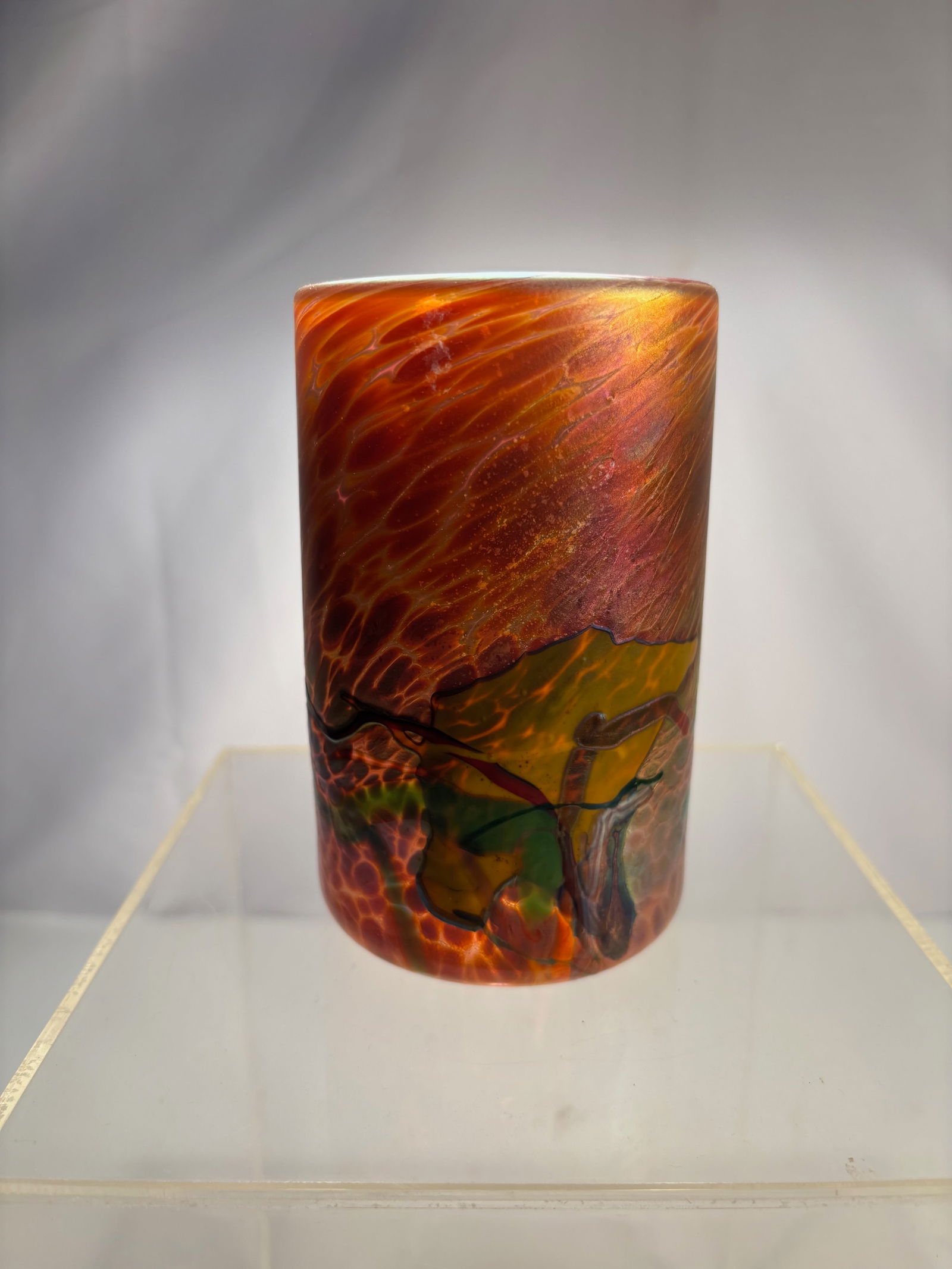 A Signed Studio Art Glass Cylinder Vase, 1999: Hand-blown studio art glass vase of tall cylindrical form, featuring richly layered molten colors in amber, orange, green, and subtle iridescent tones. The surface shows fluid, organic striations and