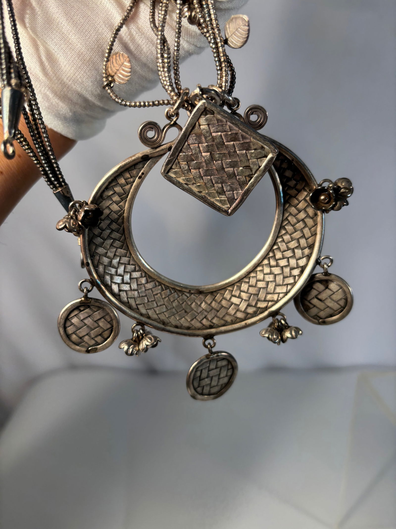 Silver Multi-Strand Necklace with Large Crescent Pendant and Decorative Disc Charms: Striking necklace featuring multiple silver chains adorned with small leaf and bead accents, suspending a large crescent-shaped pendant with woven-pattern surface and a central diamond-shaped
