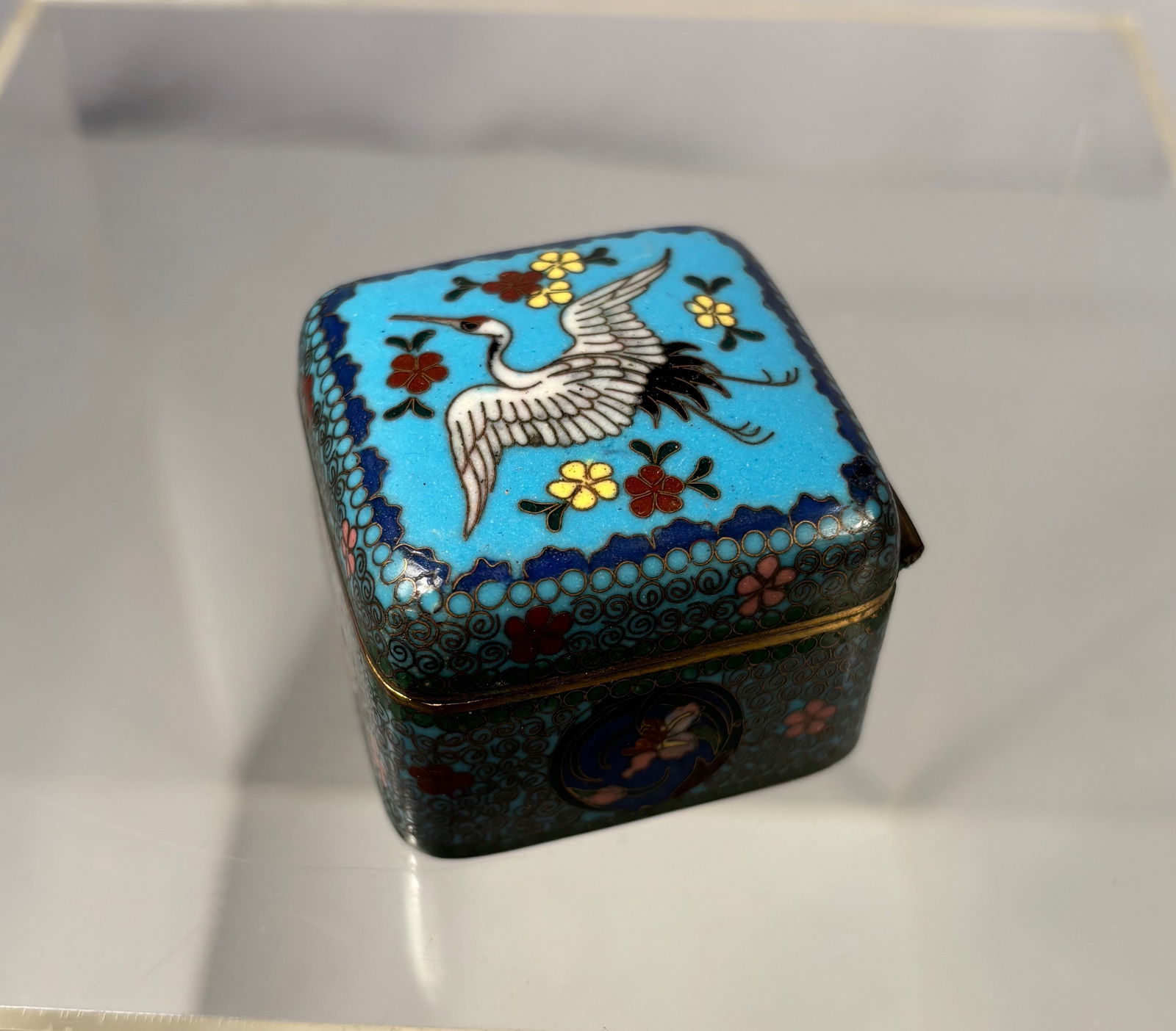 A Japanese Gilt Cloisonné Enamel Hinged Box: Meiji period (1868–1912).The square hinged box worked in cloisonné enamel on gilt metal, the cover finely decorated with a crane in flight amid floral motifs against a turquoise blue ground, the bo