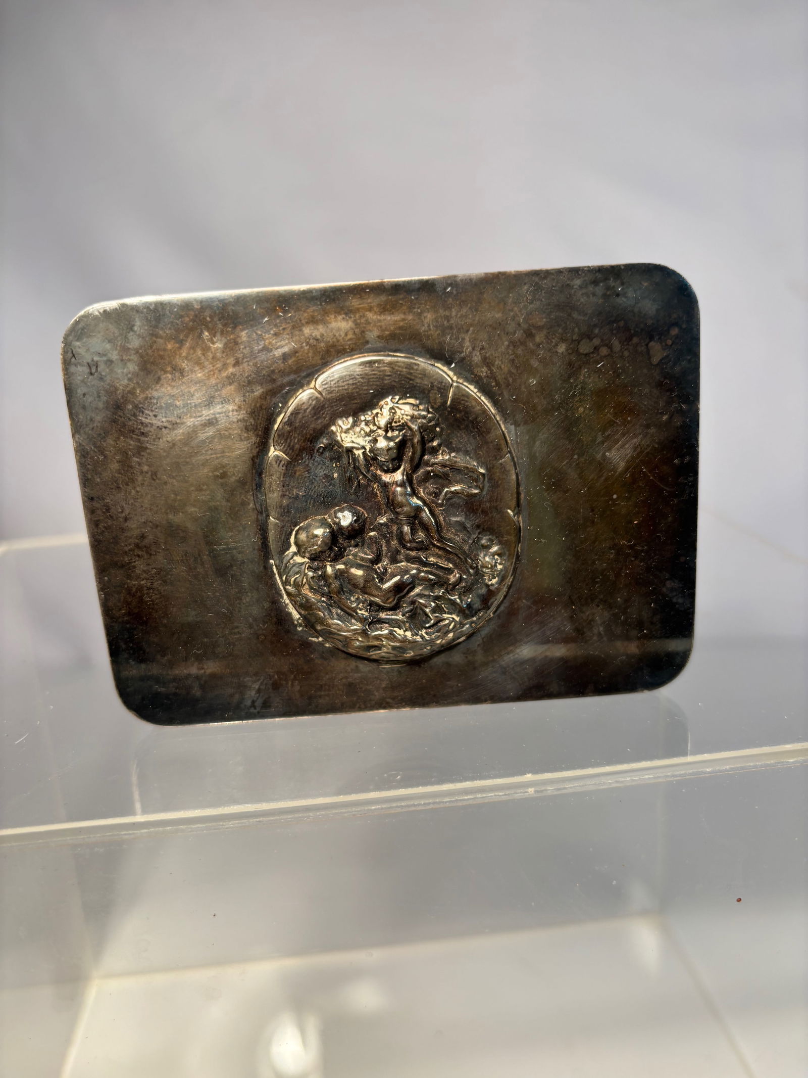 Continental Metal Card Case with Mythological Relief, Stamped “Victoria,?? Possibly Silver, circa: Rectangular continental metal card case, the front decorated with a circular relief medallion of a putto in combat with a mythological beast amid scrolling foliage. The reverse plain with age patina.