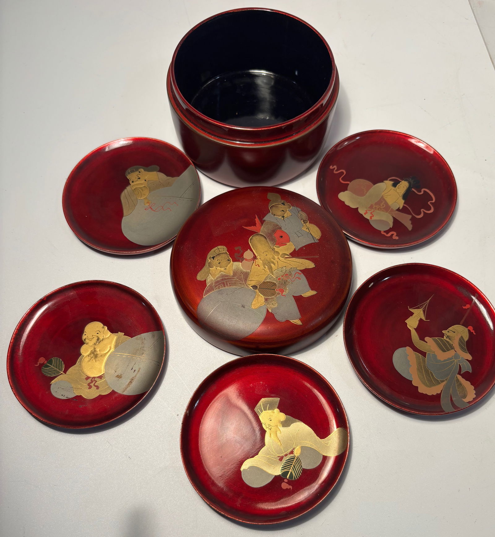 Set of Japanese Red Lacquer Box with Figural Motif and 5 Matching Dishes: 19th century, A Japanese lacquerware set comprising a circular covered box and five matching small dishes, finished in a rich red lacquer ground. The cover and dishes are decorated with gilt and color