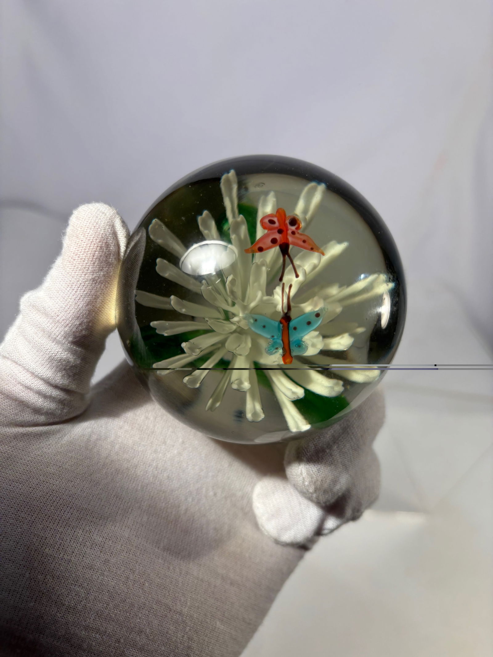 Art Glass Lampwork Floral Paperweight with Butterflies: Decorative art glass paperweight featuring a sculpted white lampwork flower rising from a green stem, set against a warm amber-toned ground. Two colorful butterflies hover above the bloom, creating a