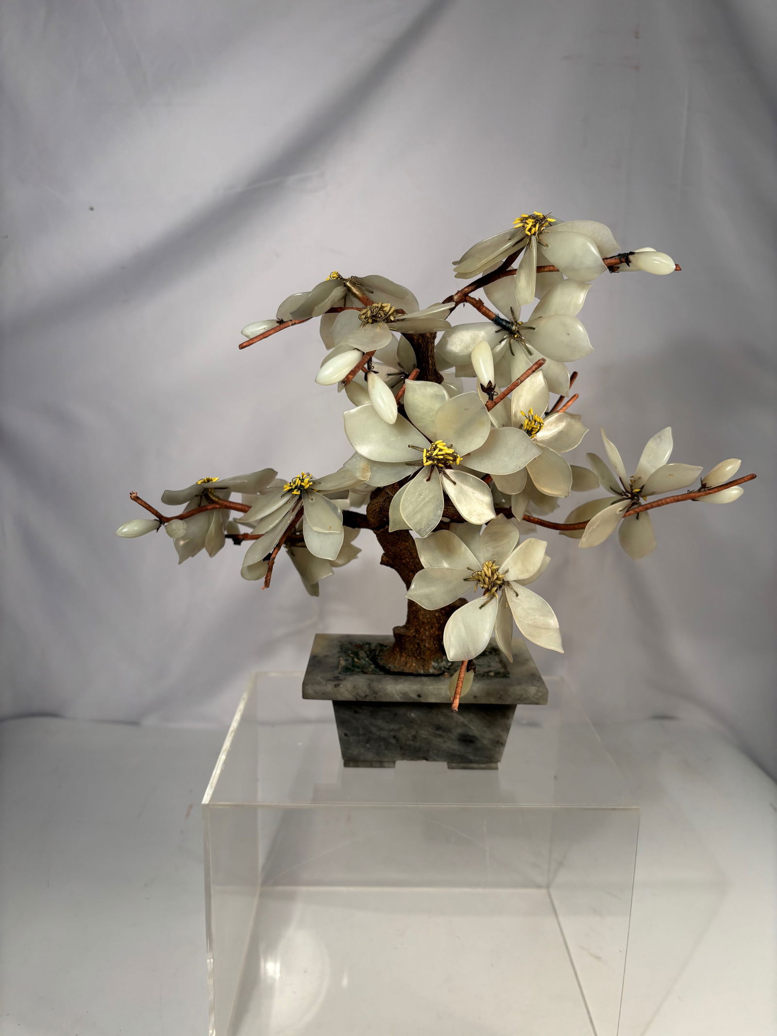 A Chinese Hardstone (Jade-Type) Blossom Bonsai Tree Sculpture: Chinese carved hardstone blossom bonsai tree, featuring pale celadon-tone stone petals arranged on wired branches over a naturalistic trunk. The sculpture is mounted on a green stone base, creating an