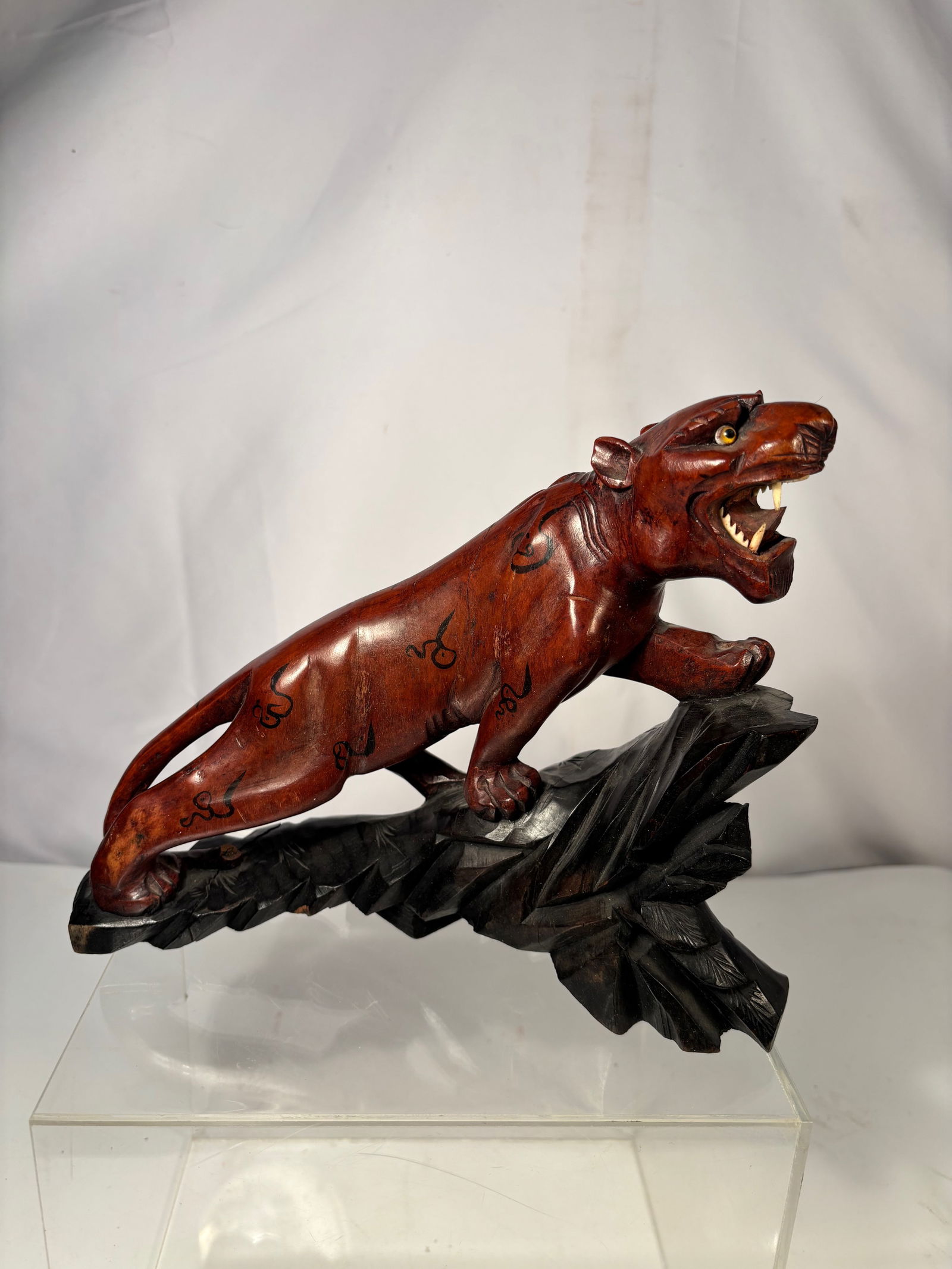 A Japanese Style Carved Tiger Figure on Wood Base: A Japanese-style carved figure depicting a tiger in a dynamic stalking pose, mounted on a carved base. The animal is rendered with expressive detail, including open jaws, incised striping, and inlaid-
